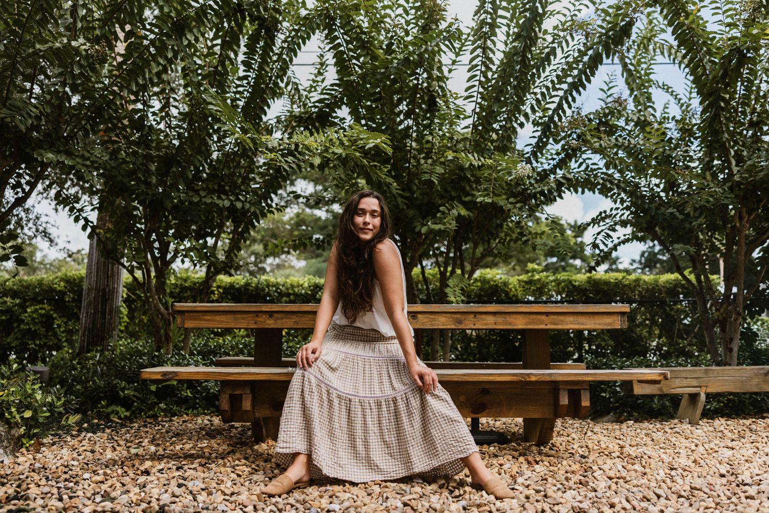 Young woman in a linen tiered maxi skirt sitting on a wooden bench surrounded by lush green trees.