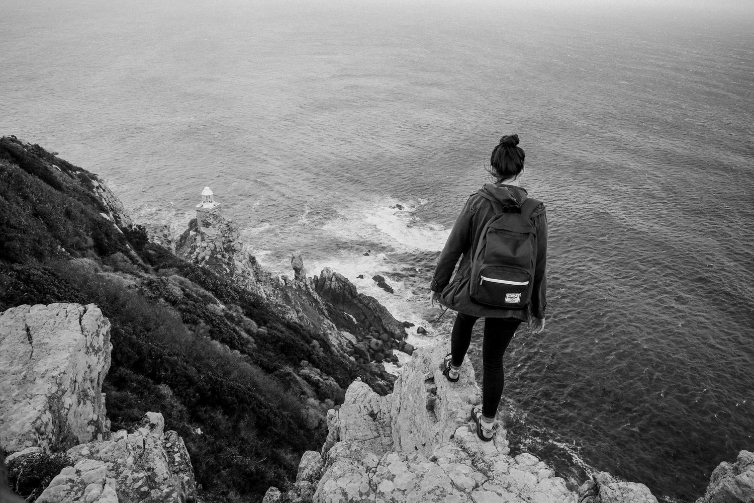 Black and white photo of a hiker with backpack standing on a rocky cliff overlooking the ocean and a lighthouse.