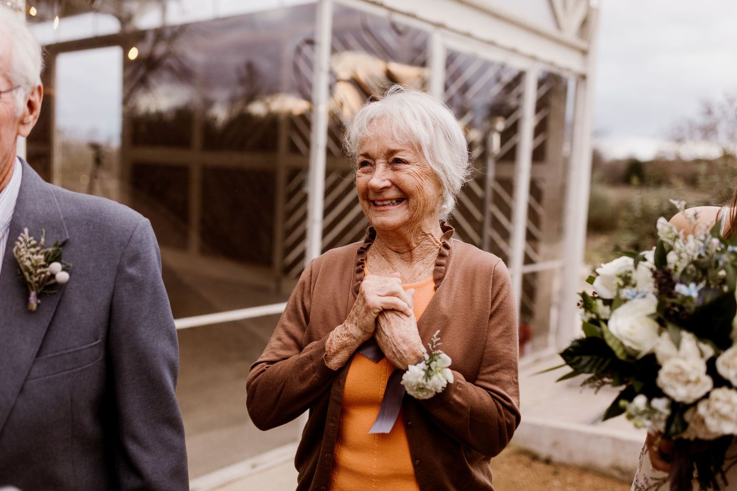 Elderly woman smiling joyfully with clasped hands wearing a wrist corsage at an outdoor wedding ceremony.