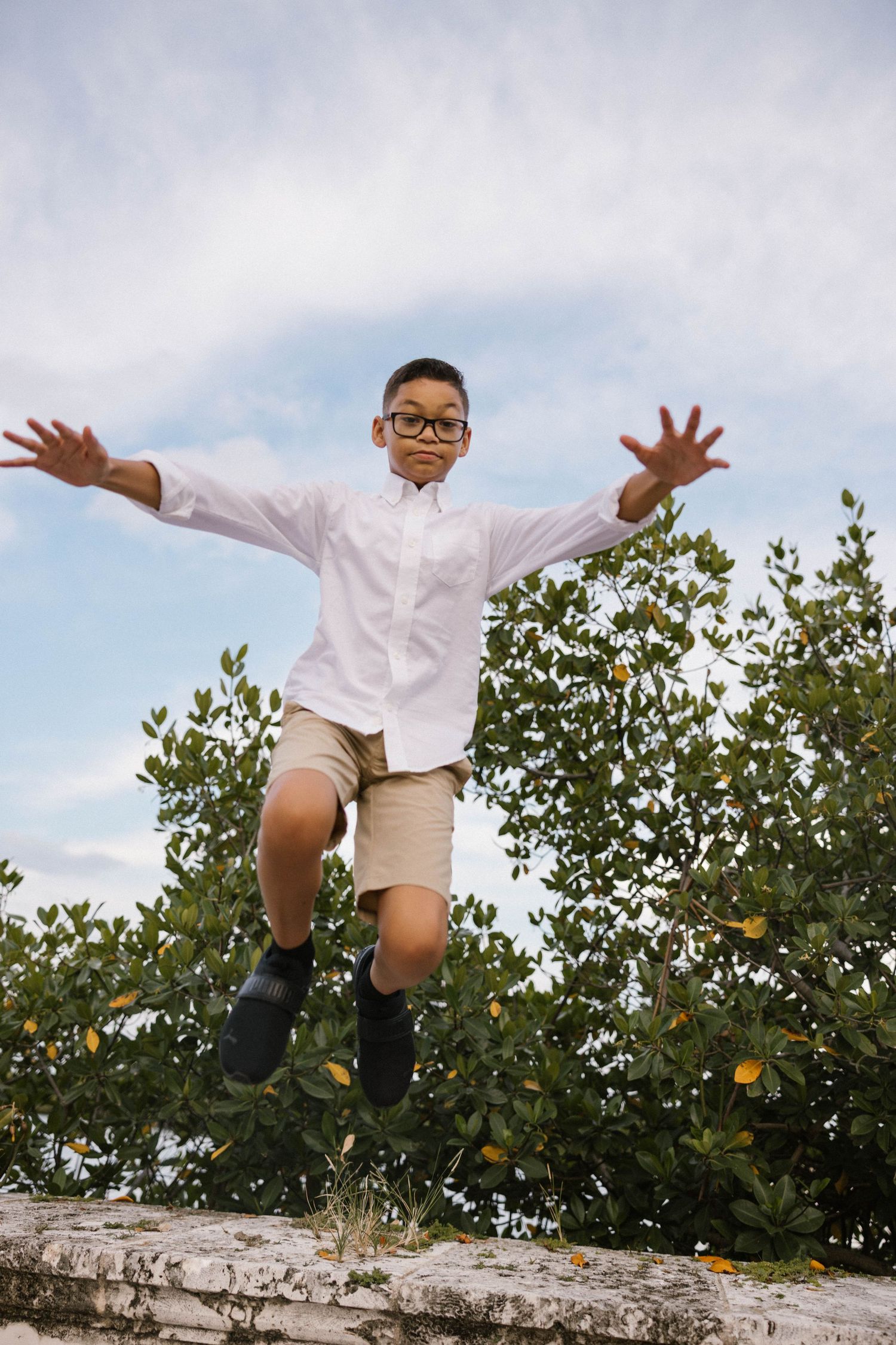 Young boy in white shirt and khaki shorts jumps off a stone wall with arms outstretched.