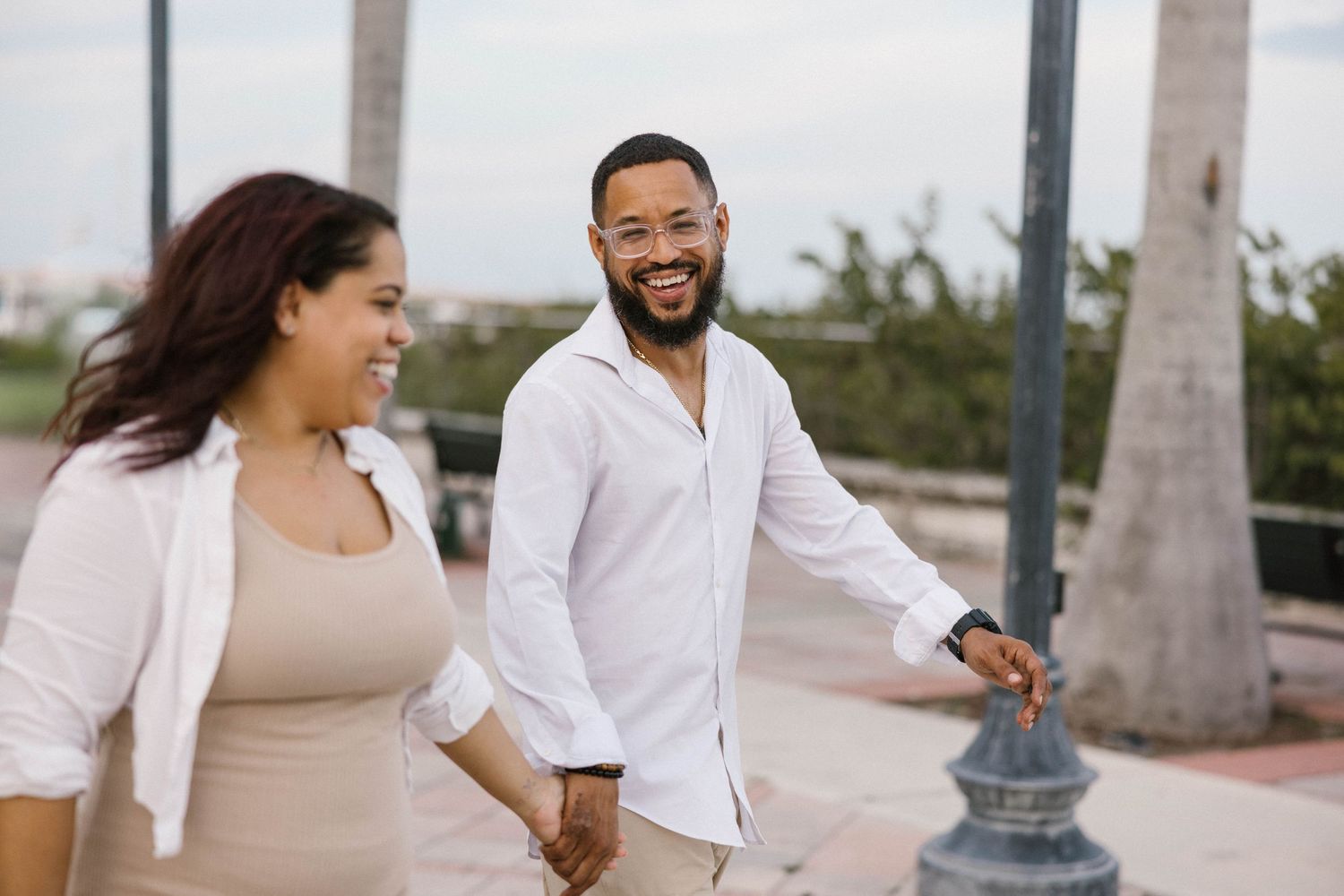 Smiling man in a white shirt holds hands with a woman while walking outdoors near palm trees.