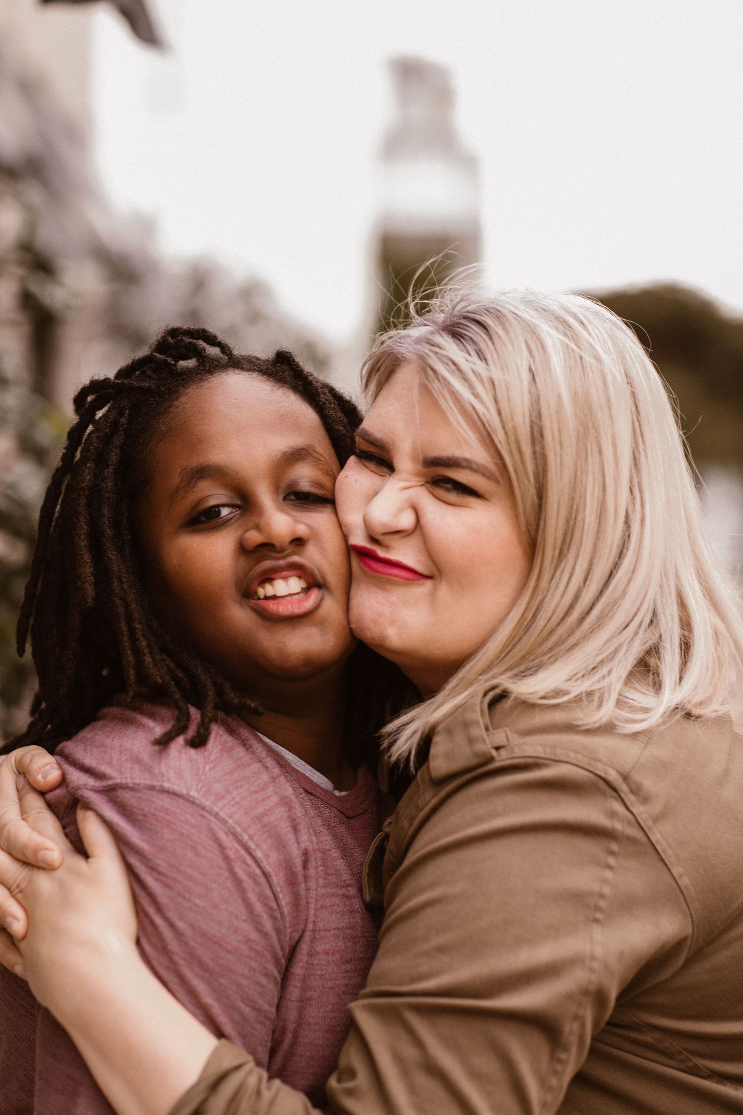 Blonde woman kissing the cheek of a smiling child with locs in a warm outdoor embrace.