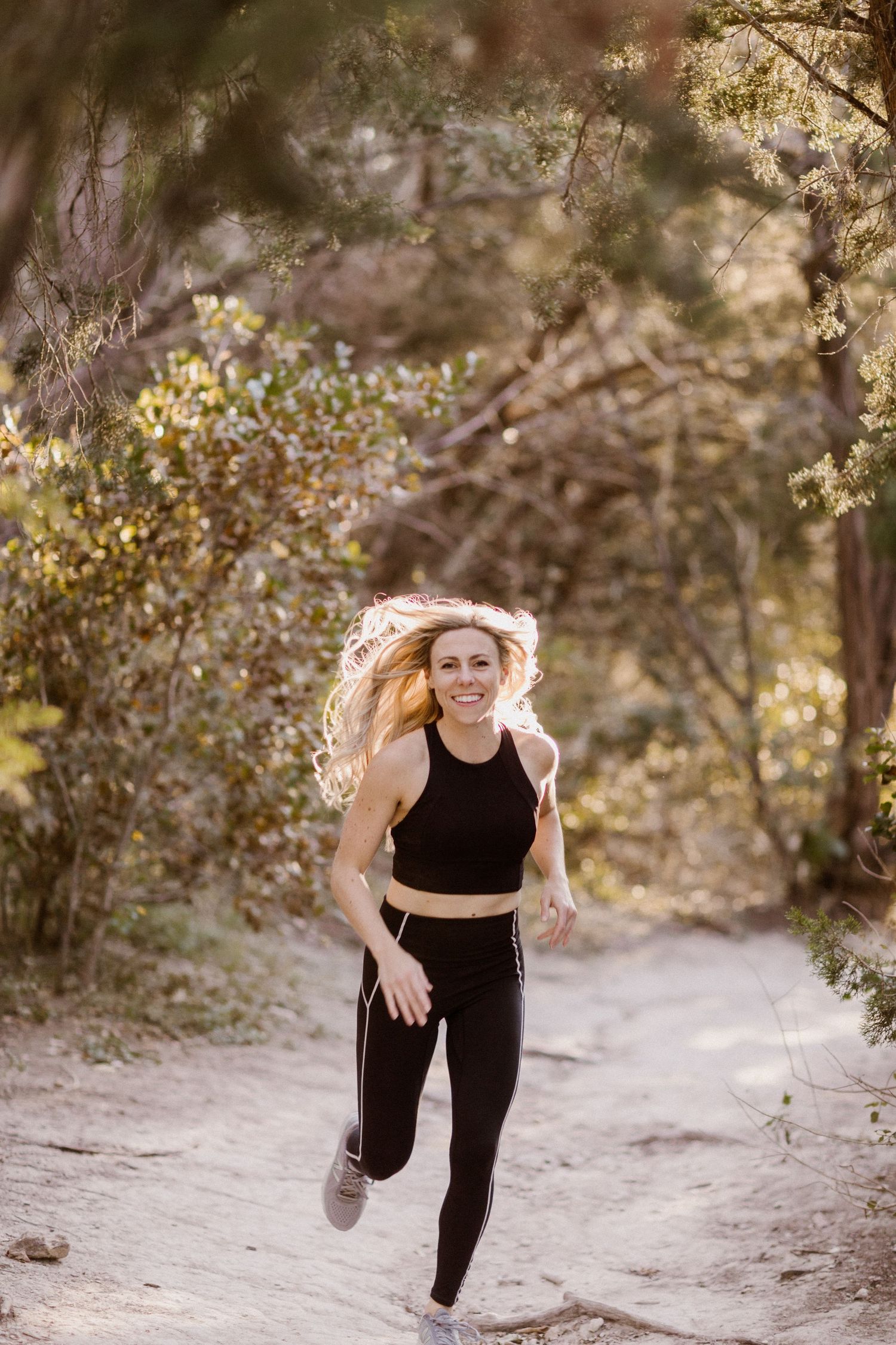 Smiling woman with blonde hair running on a forest trail wearing black athletic crop top and leggings.