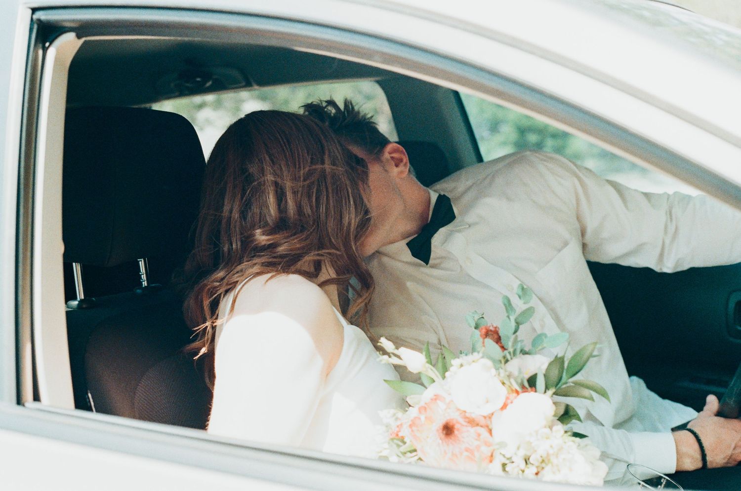 Newlywed couple sharing a romantic kiss inside a car, bride holding a pastel bouquet of pink and white flowers.