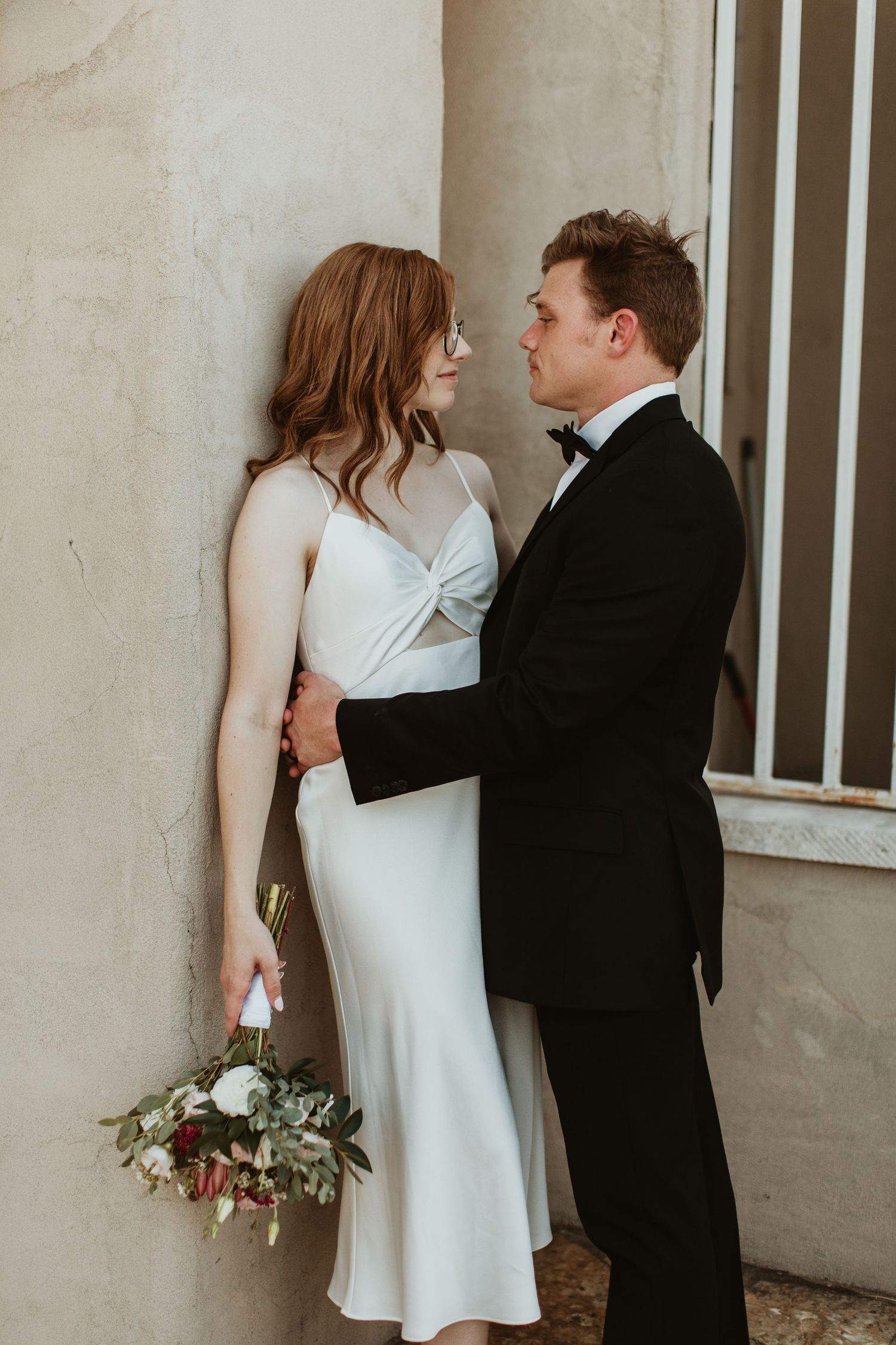 Bride in white spaghetti strap dress holding bouquet leans against wall with groom in black tuxedo.