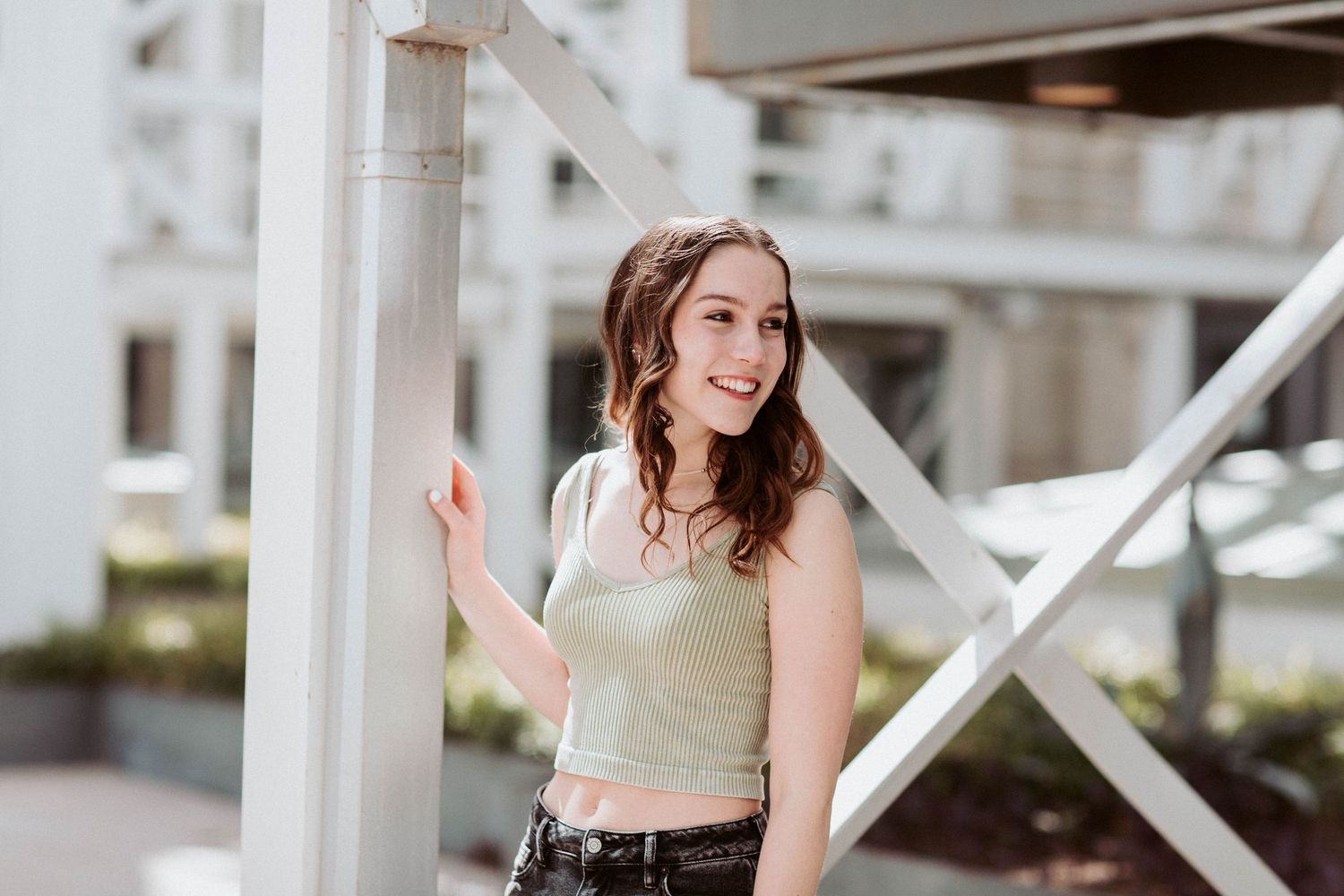 Smiling young woman in mint crop top leans against a white pillar in a bright outdoor setting.