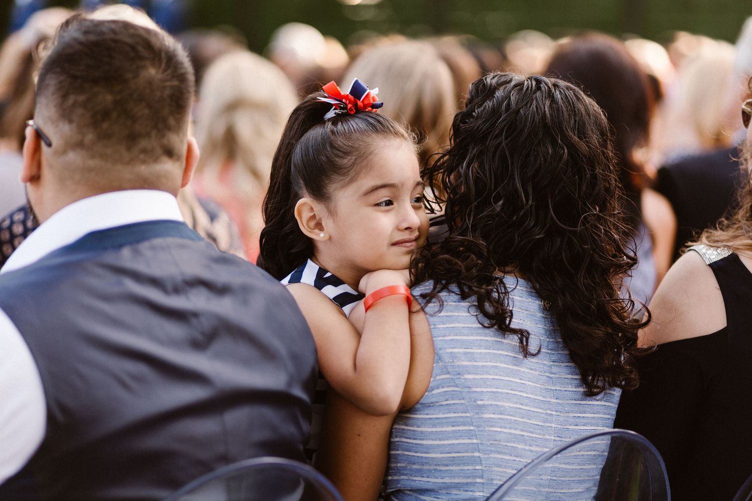 Young girl with a red bow sits on an adults lap during an outdoor wedding ceremony.