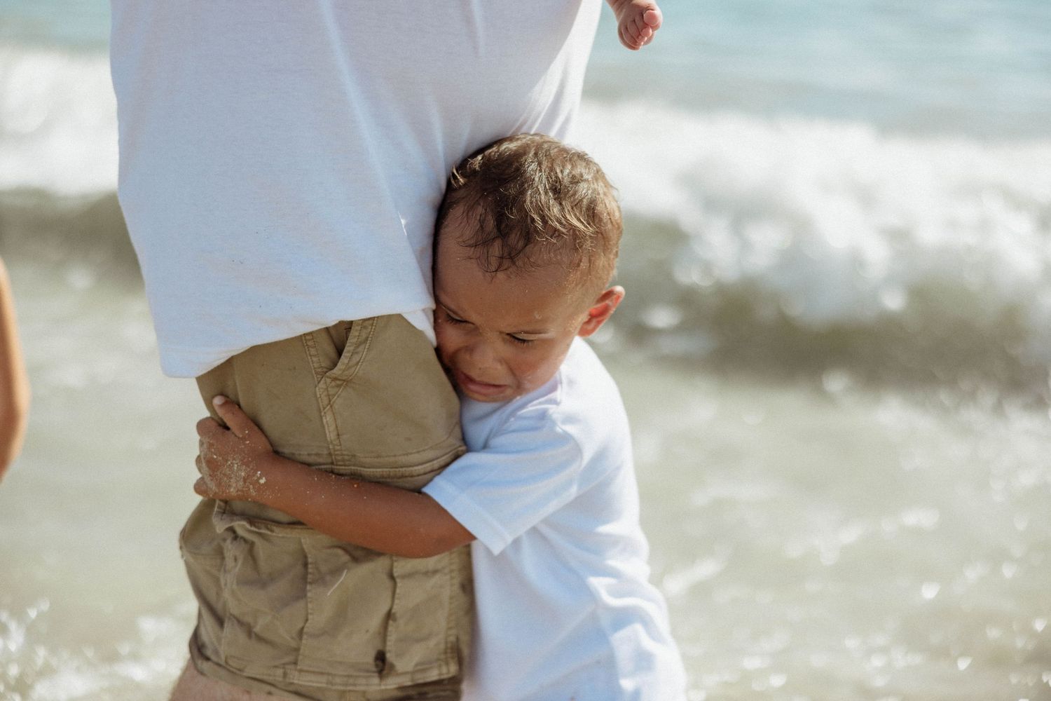 Young boy hugging an adult tightly on a sunny beach with ocean waves behind them.