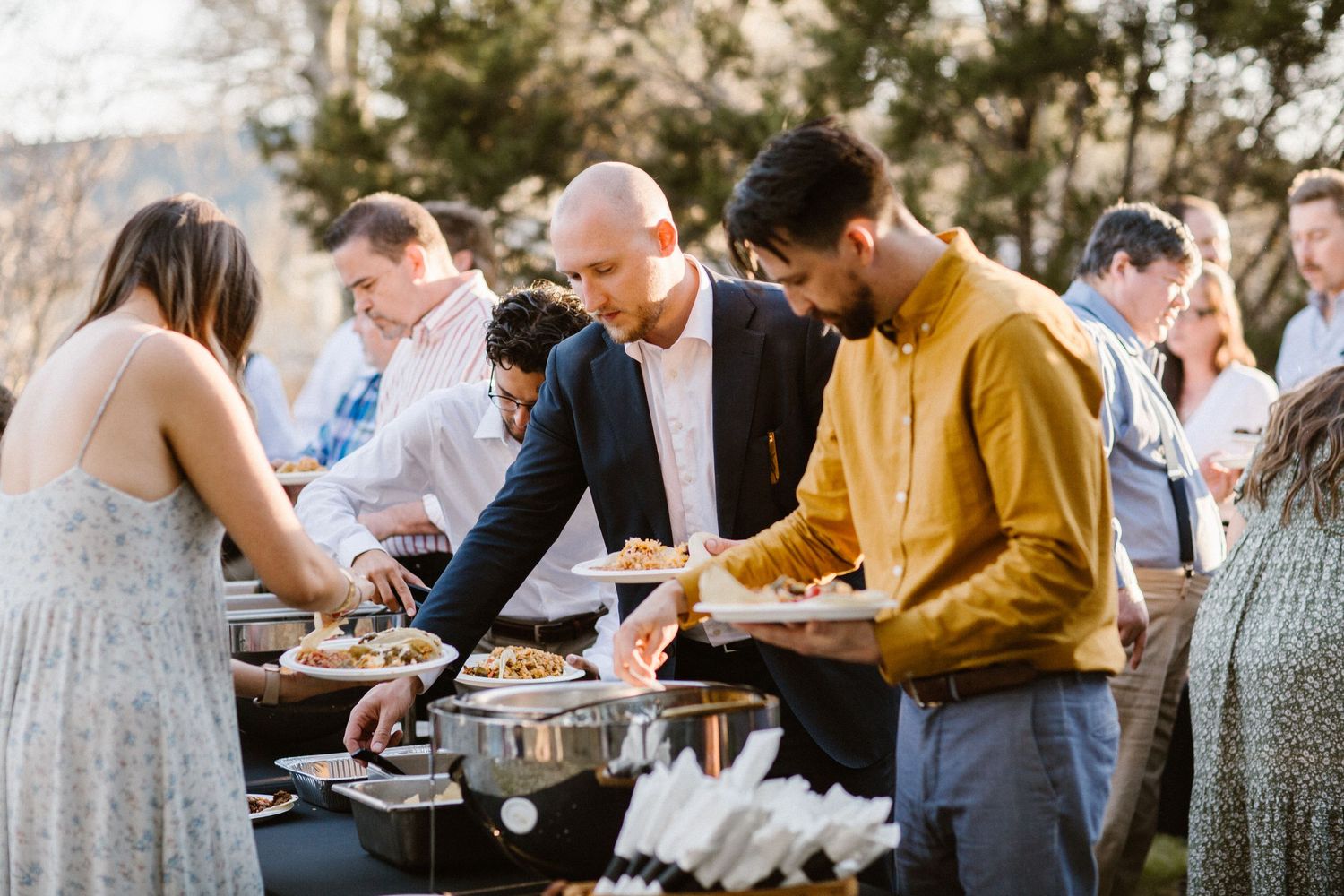 Wedding guests serve themselves from a buffet line at an outdoor catered reception.