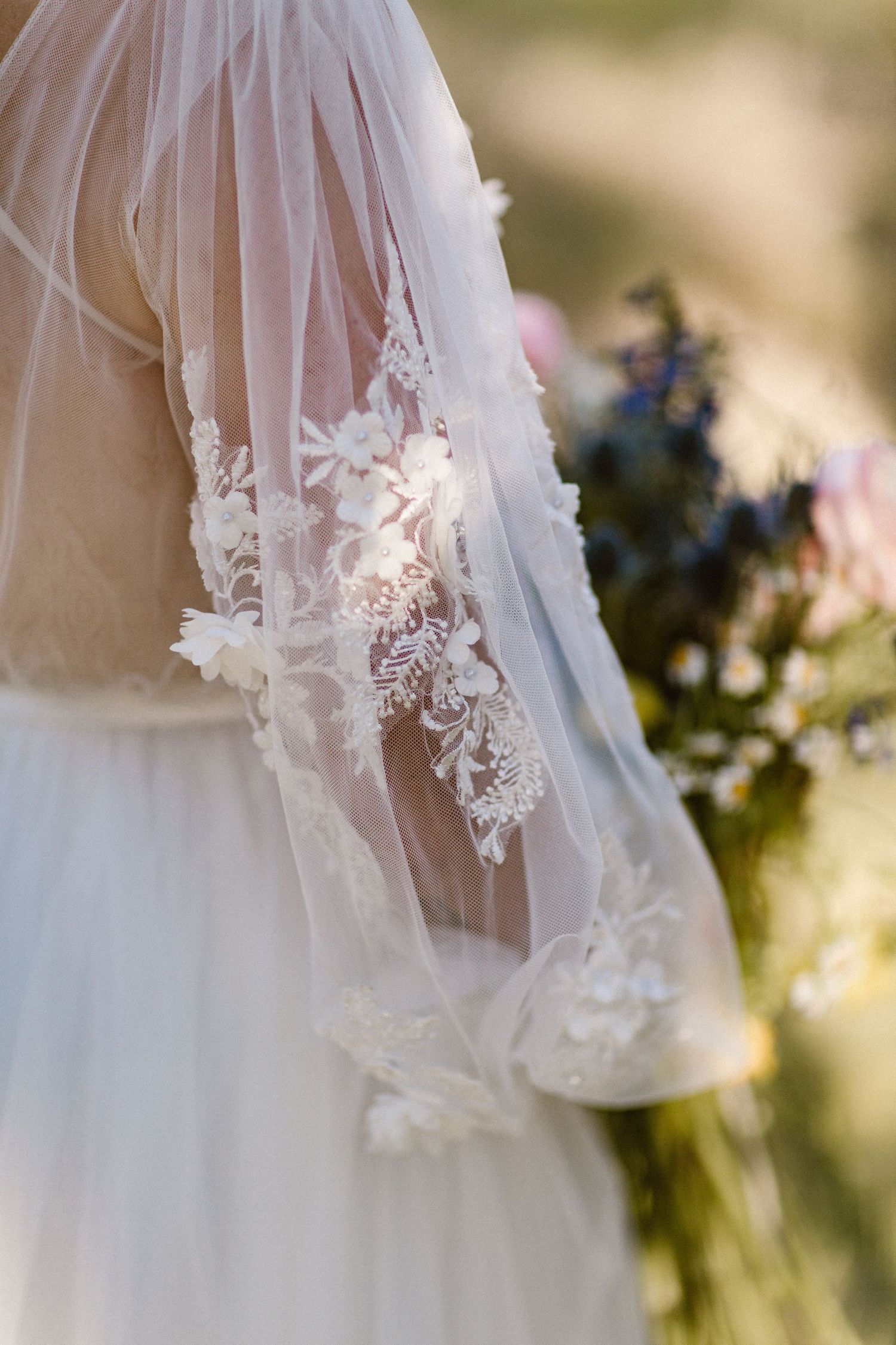 Detail shot of bride's sheer lace wedding dress sleeve with intricate floral embroidery in sunlight.