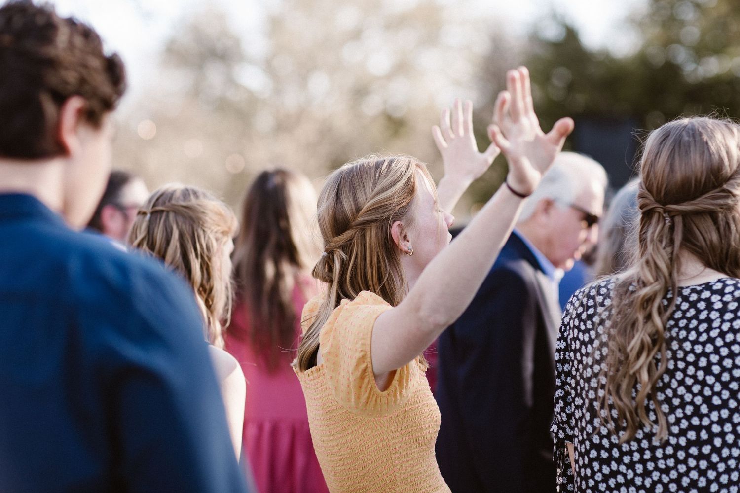 Wedding guest in yellow dress raises hand in worship during outdoor ceremony among other guests.