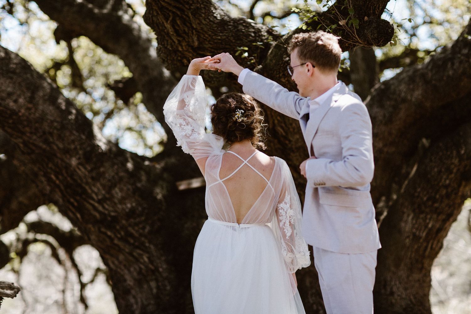 Groom twirling bride in a stunning backless lace wedding gown beneath a sprawling oak tree.