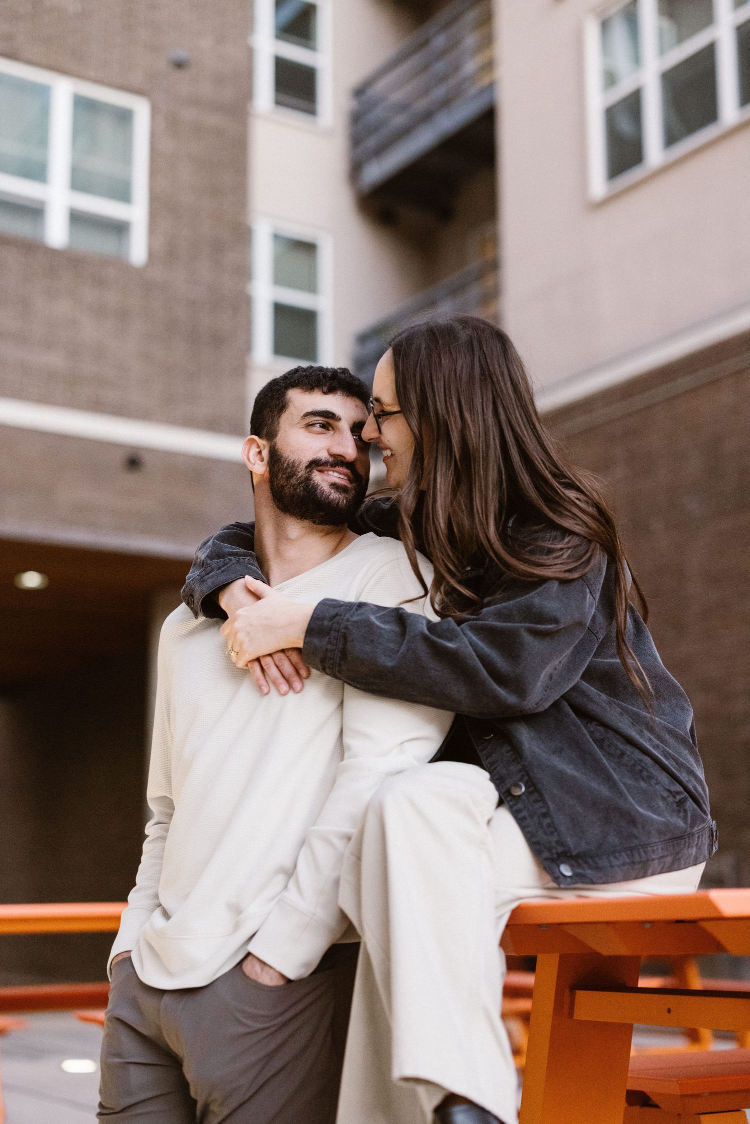 Woman embracing man from behind while sitting on an orange bench outdoors.
