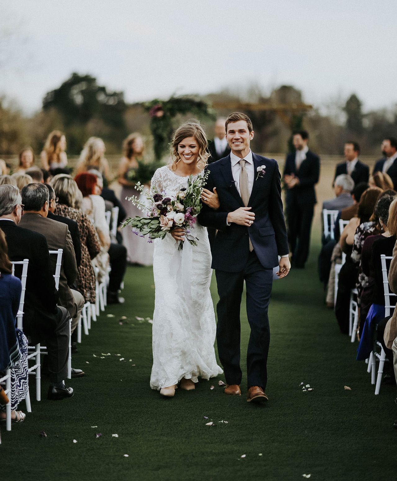 Smiling bride and groom walk down outdoor wedding aisle lined with seated guests and white chairs.
