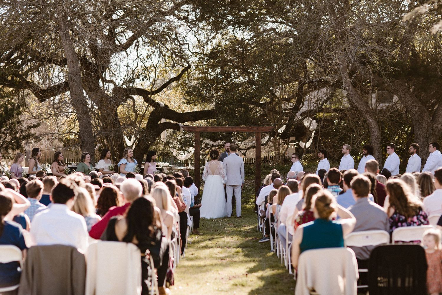 Outdoor wedding ceremony with guests seated in white chairs under large oak trees.