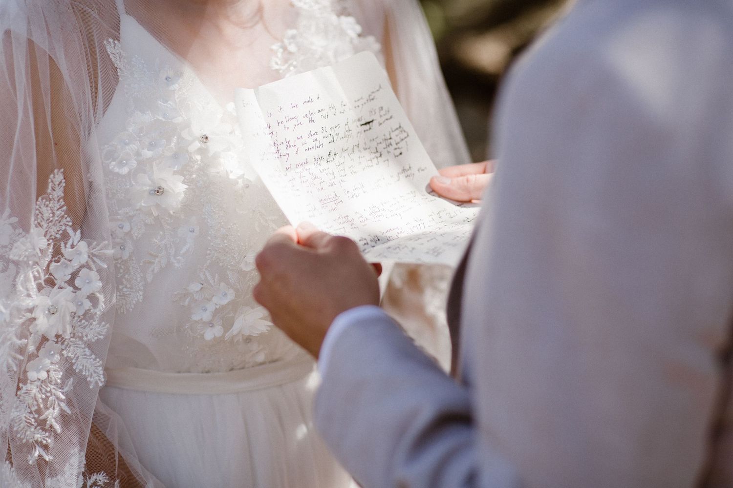 Groom reads handwritten vows while holding brides lace-gloved hands during outdoor wedding ceremony.