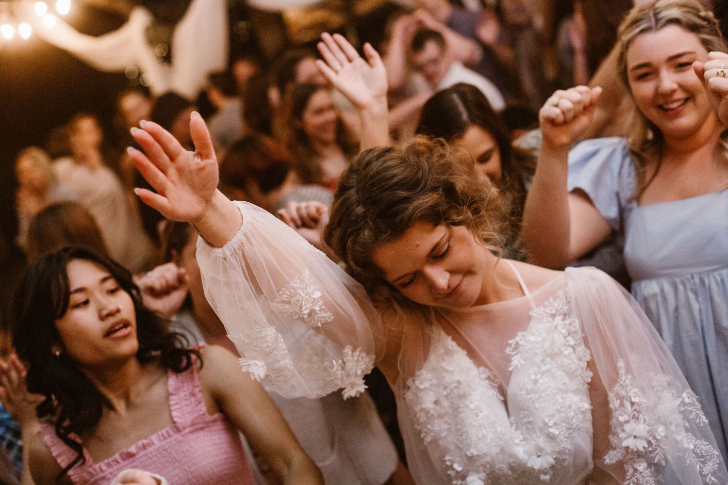 Bride in sheer lace gown dancing energetically with guests at a lively wedding reception.