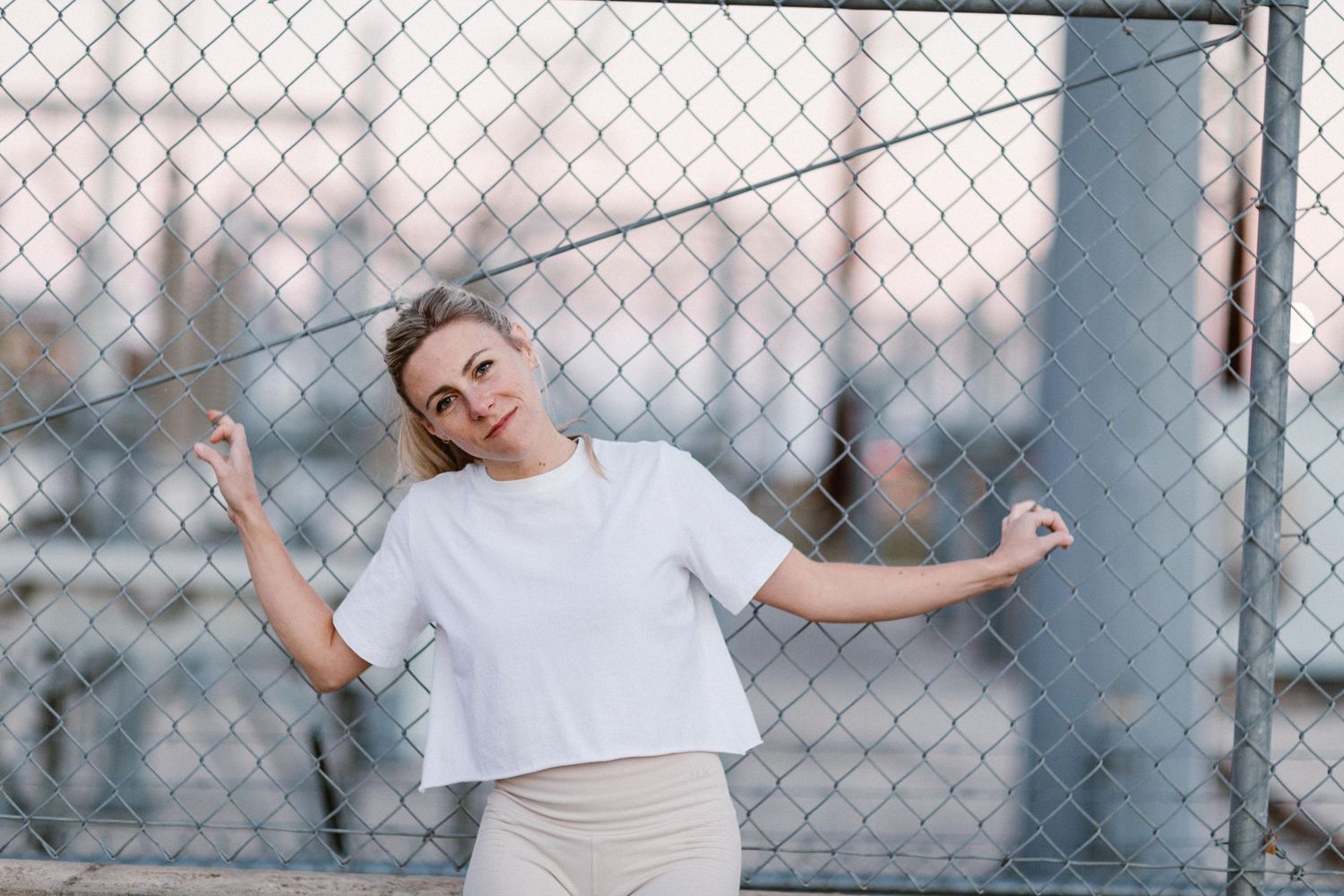 Young blonde woman in white crop top leaning against chain-link fence outdoors at dusk.