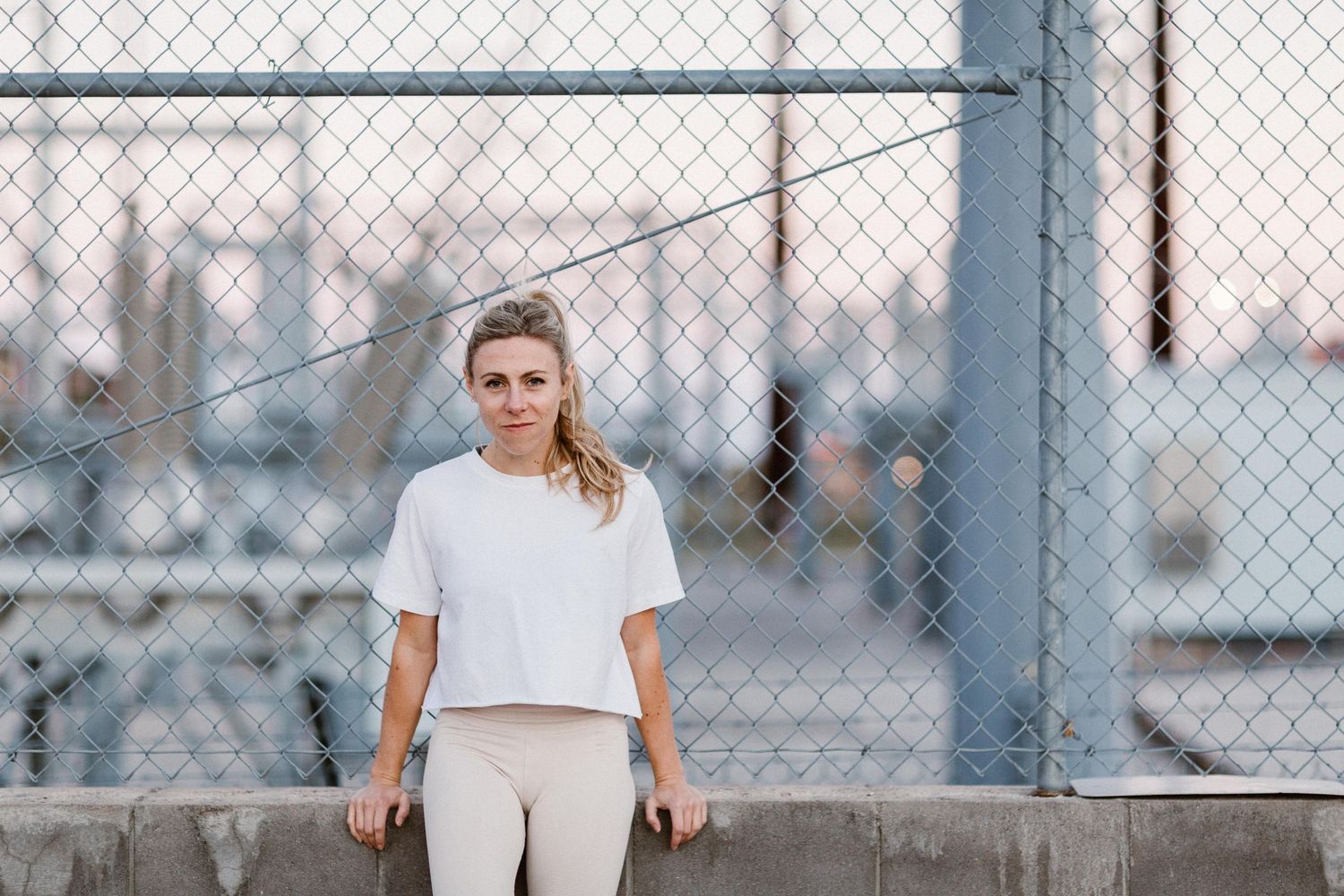 Woman in white crop top and beige leggings leaning against concrete wall by chain-link fence near marina.
