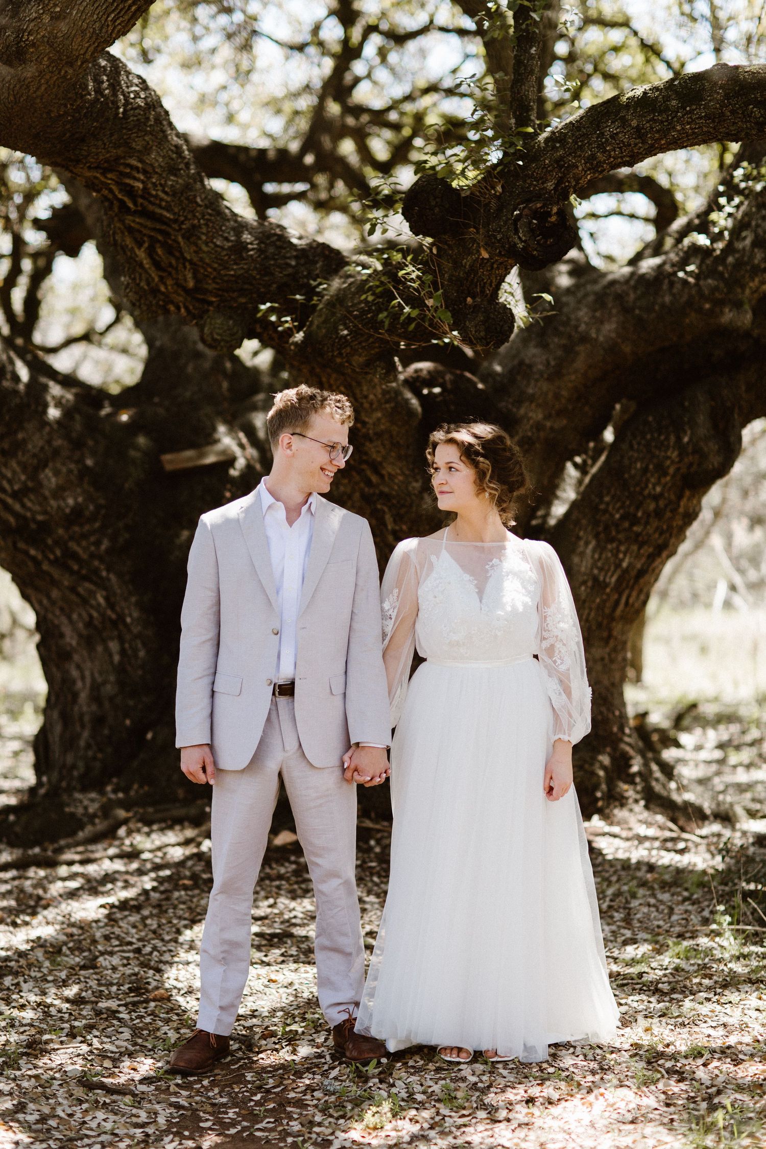 Bride in white gown and groom in tan suit hold hands beneath ancient oak tree.