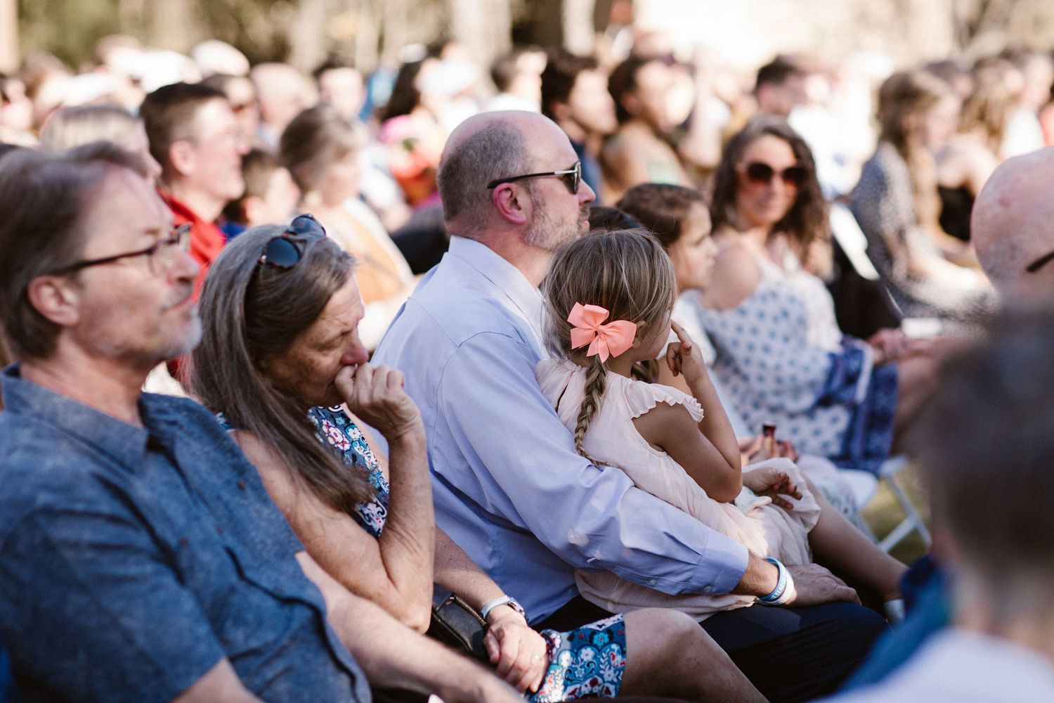 Family with young girl wearing a pink flower sits together at an outdoor wedding ceremony.