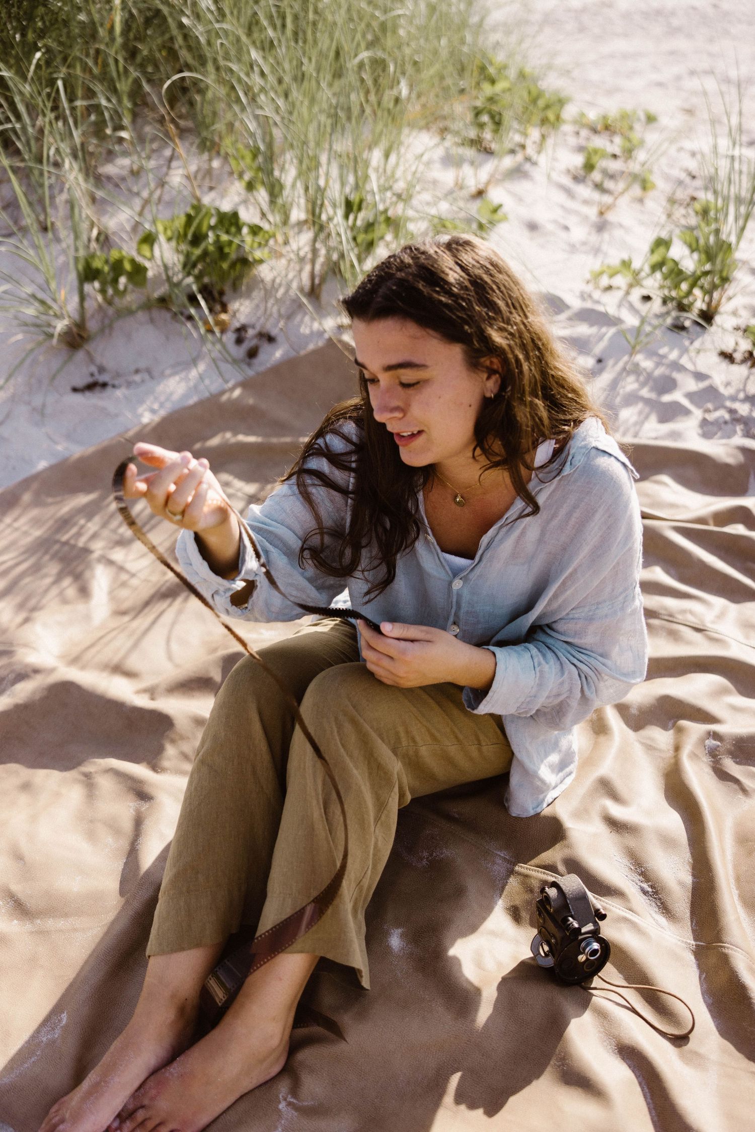 Woman sitting on beach blanket examining camera film strip in sunny sand dunes.