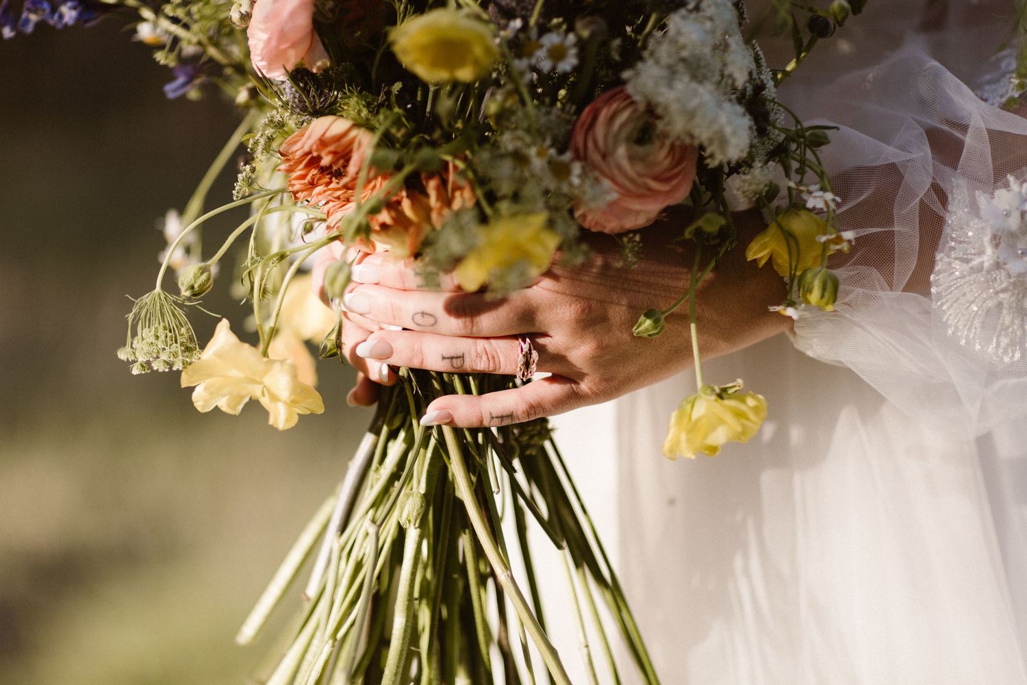 Bride holding a lush wildflower bouquet with pink, yellow, and white blooms tied with pink ribbon.
