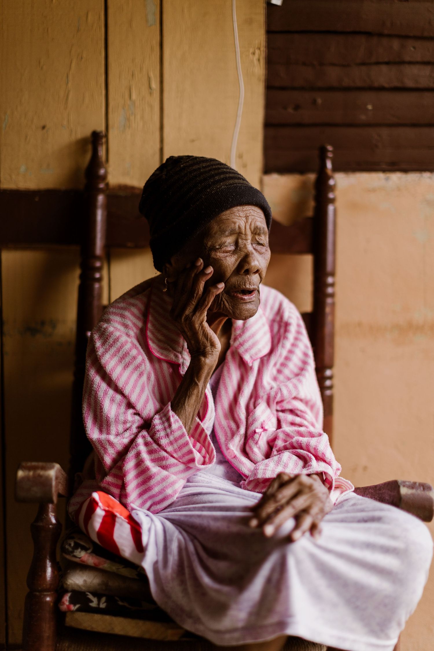 An elderly woman in a pink striped robe sits in a wooden chair talking on a mobile phone.