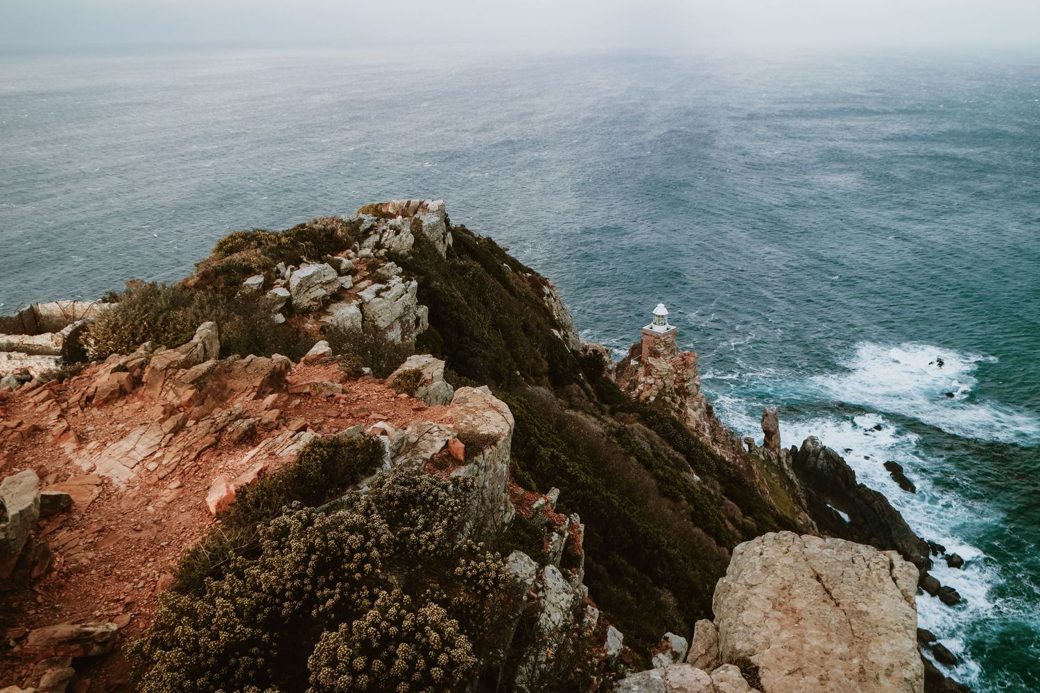 Rocky coastal cliff covered in green shrubs with a white lighthouse overlooking a choppy grey ocean.