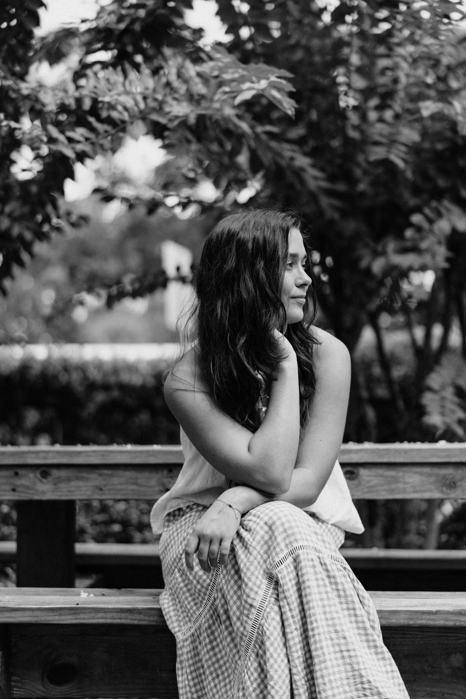 Black and white photo of a young woman in a plaid skirt sitting on a wooden bench outdoors.