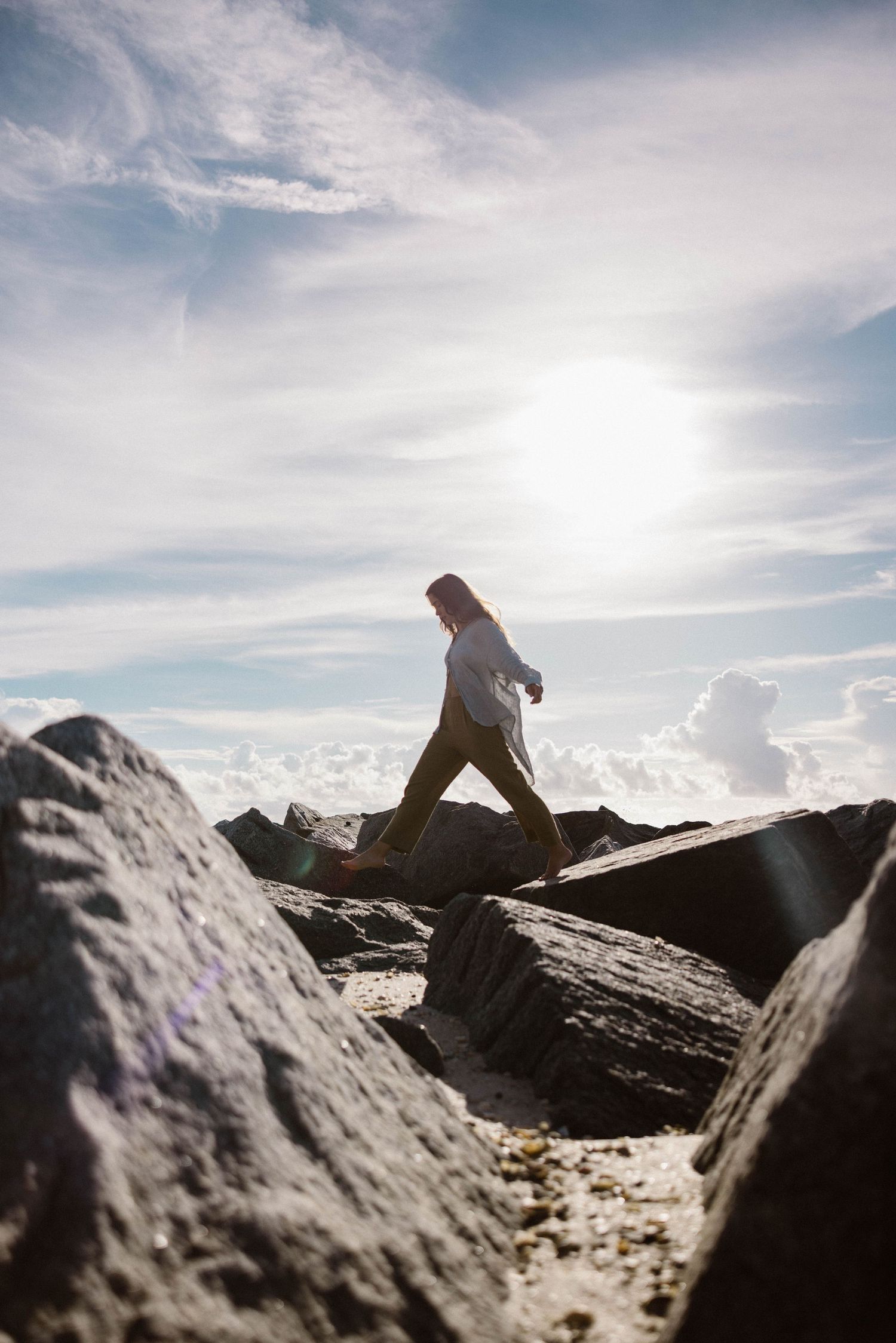 Silhouette of a woman walking across rocks against a bright sunny sky at the beach.