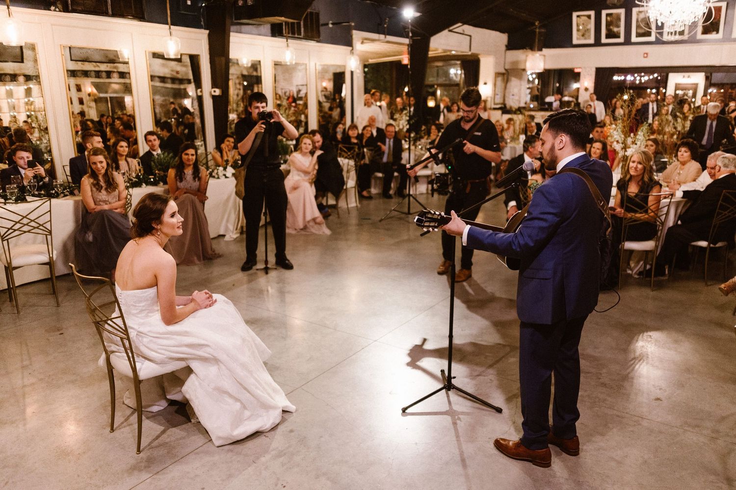 Bride seated as groom performs a heartfelt song on guitar at indoor wedding reception.
