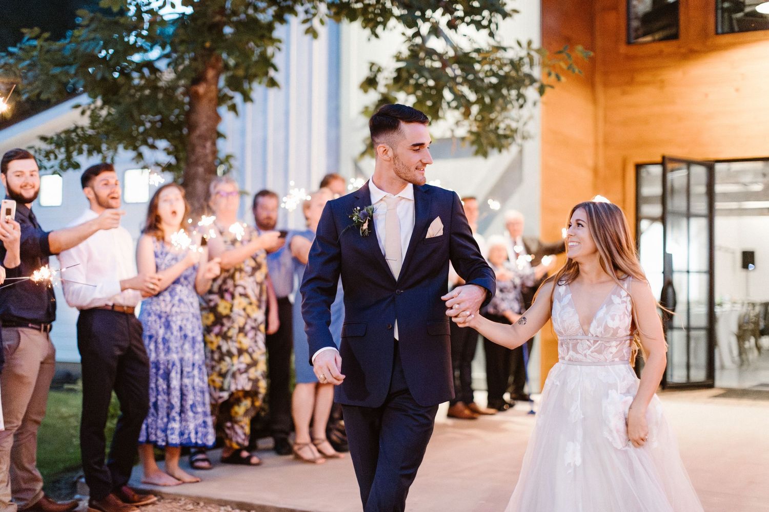 Bride and groom walking hand in hand through a sparkler send-off surrounded by cheering wedding guests at dusk.
