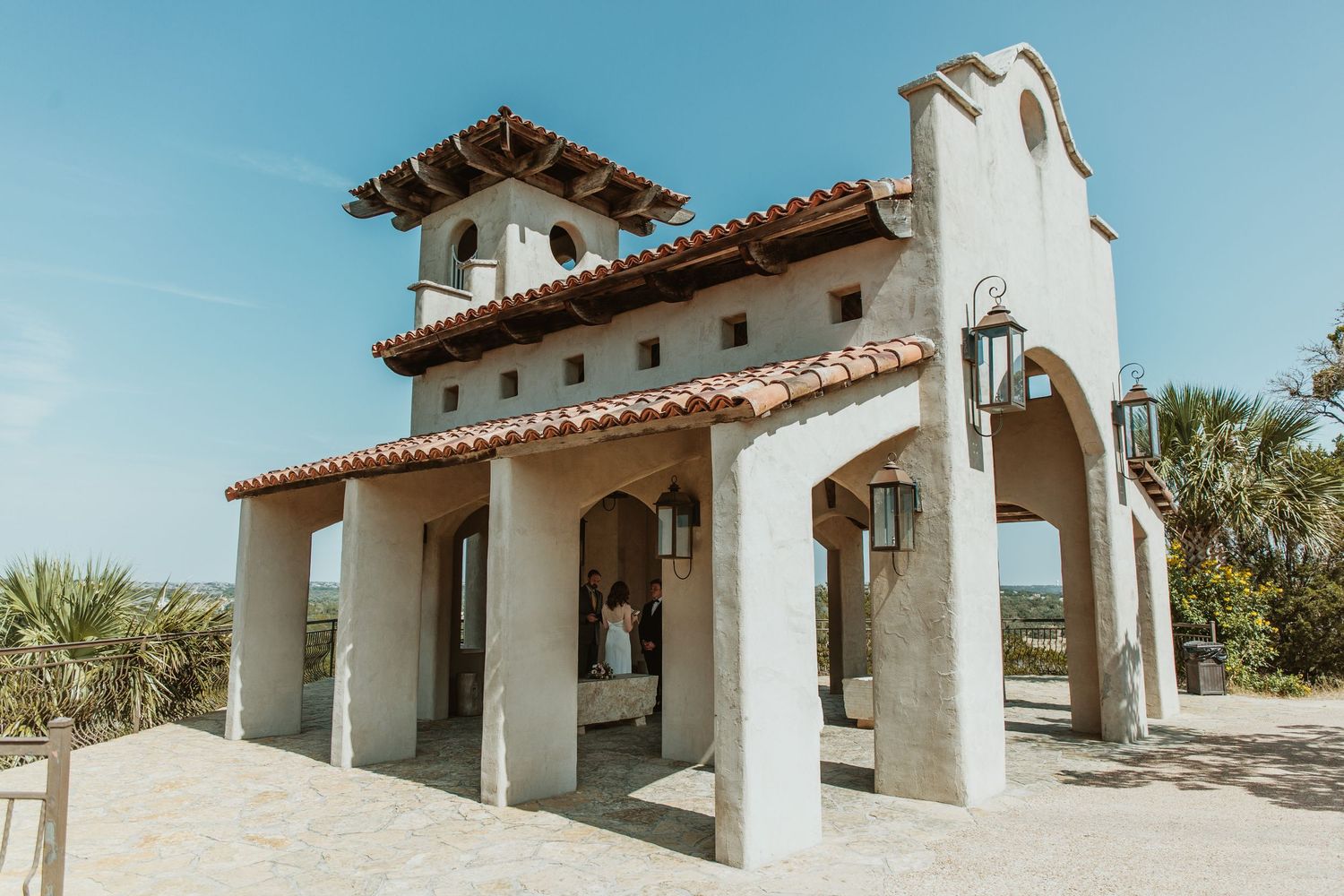 Spanish Colonial Revival chapel with red tile roof and bell tower under blue sky, perfect for outdoor weddings.