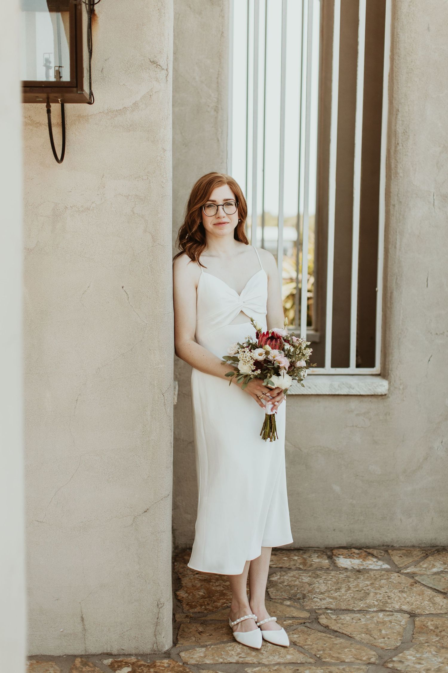 Bride in white midi slip dress holding bouquet, standing by stone wall in white heels and glasses.
