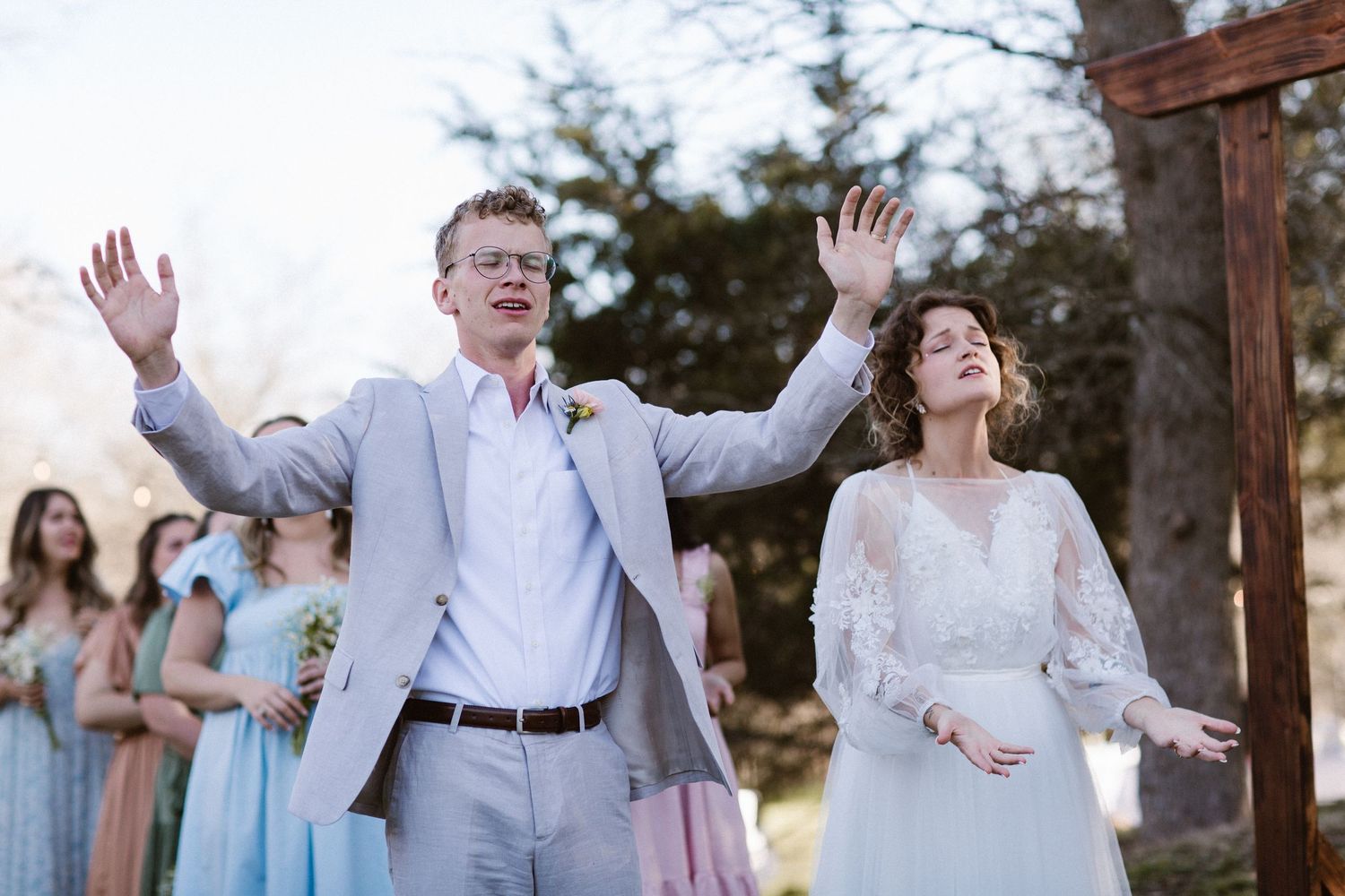 Joyful bride and groom raising hands in celebration at outdoor wedding ceremony with bridesmaids in blue dresses.