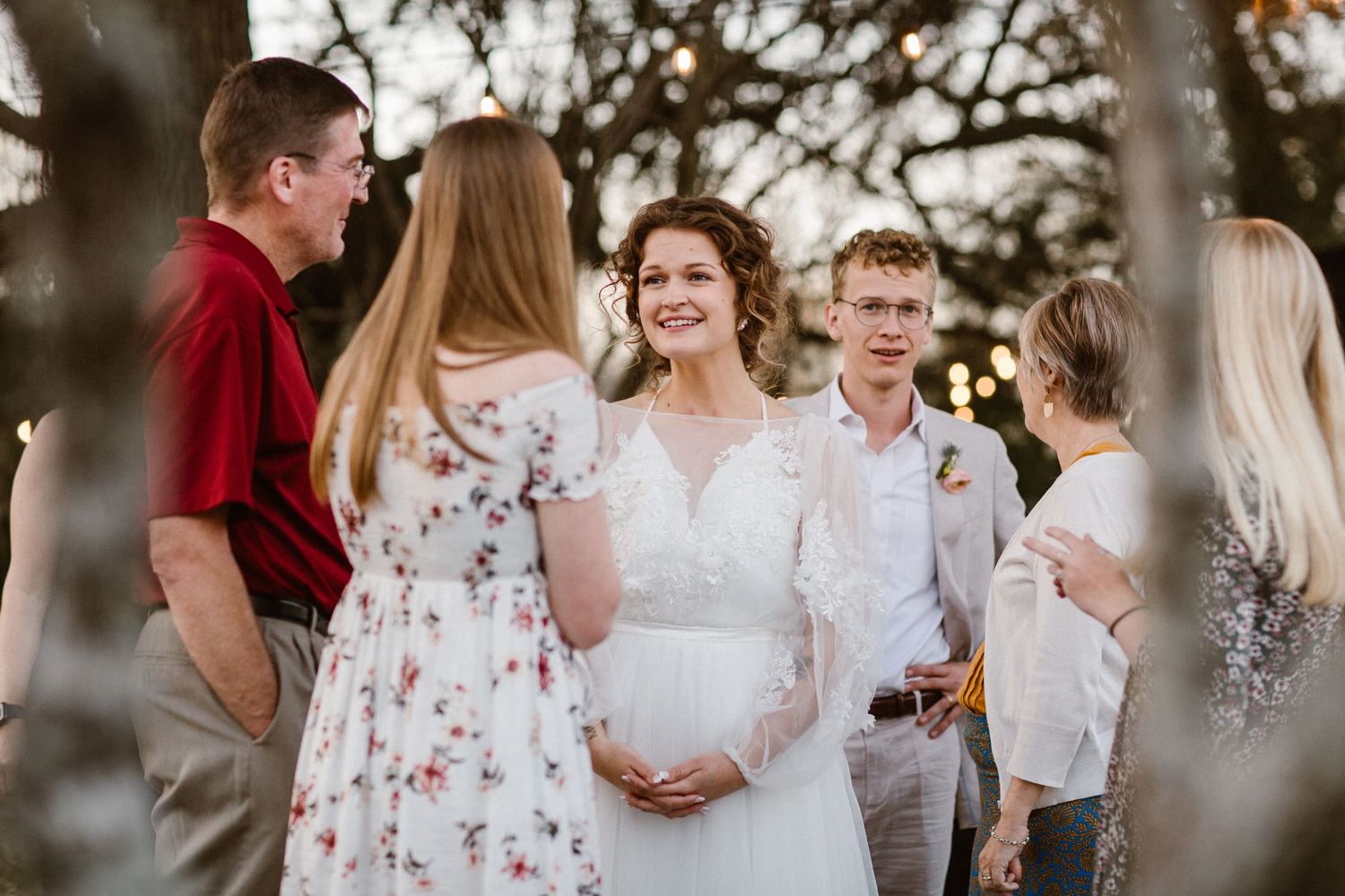 A smiling bride in a lace gown greets guests among trees at her outdoor wedding.