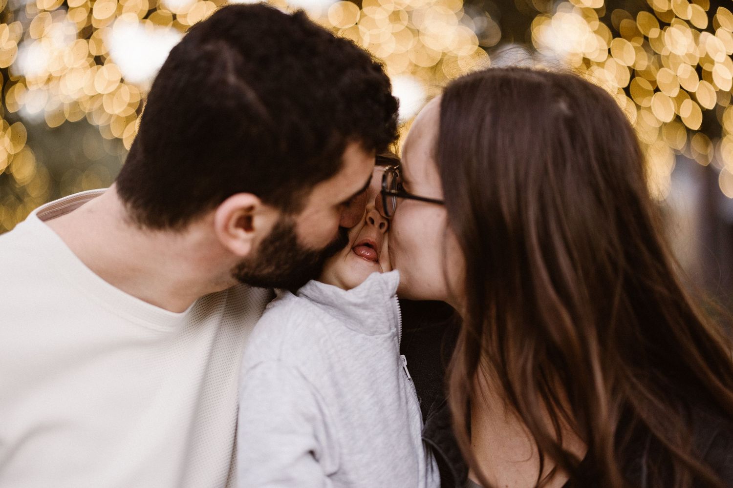 Couple sharing a tender moment kissing their smiling child with bokeh lights behind.