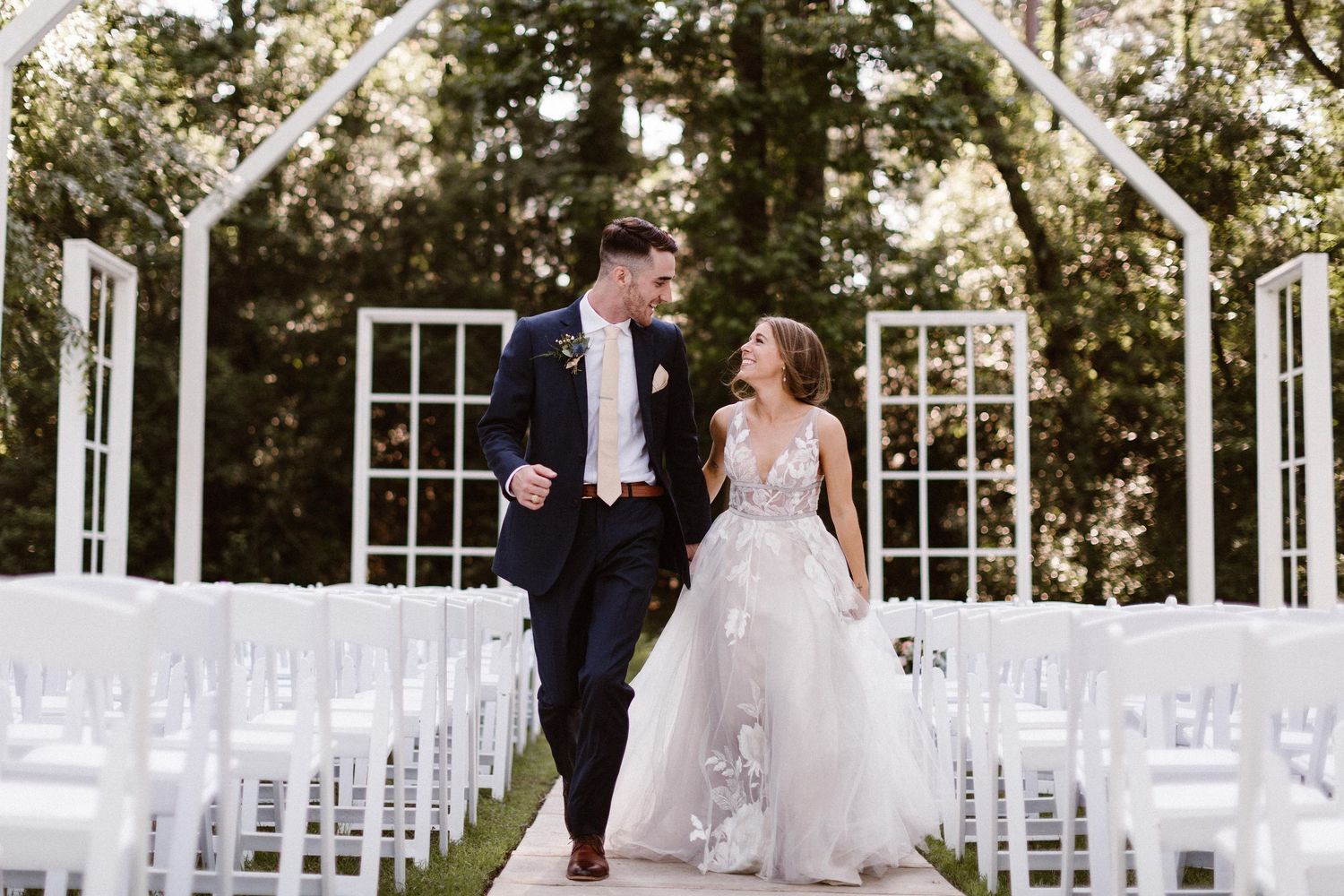 Bride and groom walking joyfully down outdoor aisle with white chairs and arch backdrop.