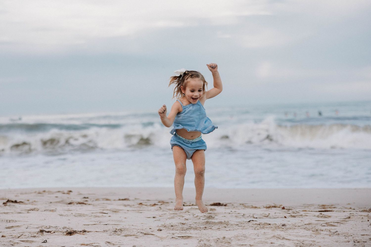 Happy toddler girl in blue outfit running playfully along a sandy beach with waves behind her.