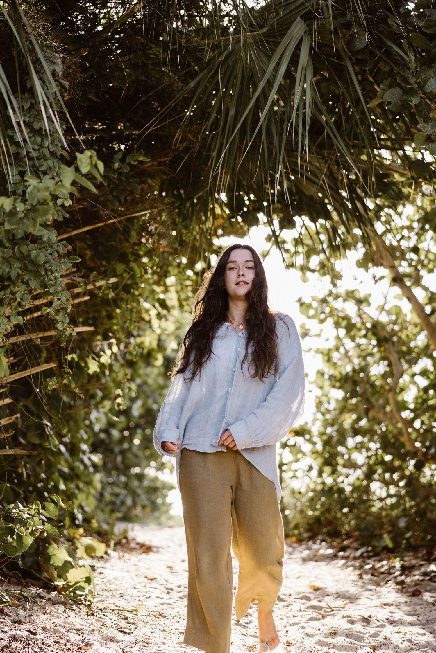 Young woman in olive linen pants and light blue shirt standing on a sandy path in tropical foliage.