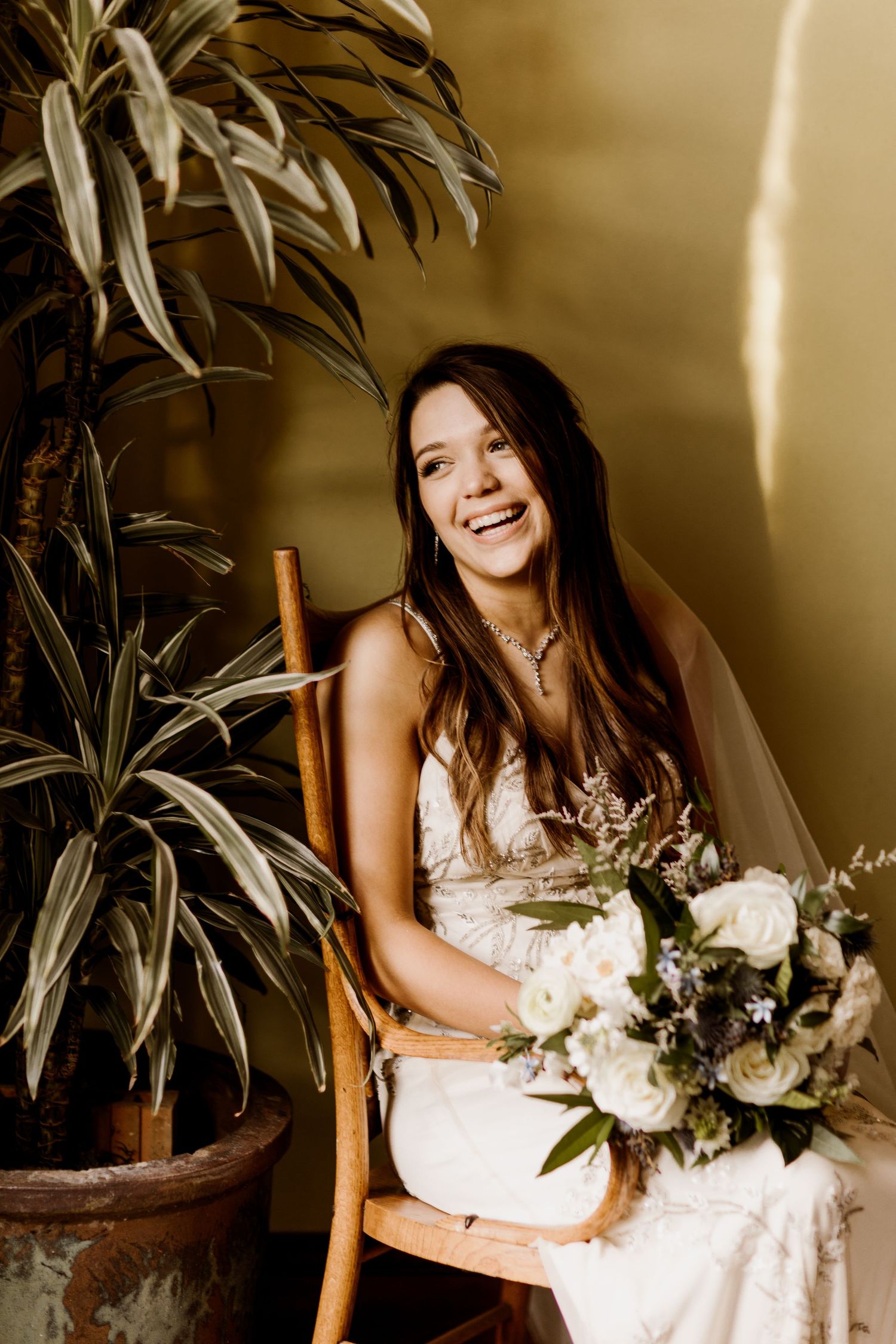 Radiant bride in white gown sitting in wooden chair holding white floral bouquet next to tropical plant.