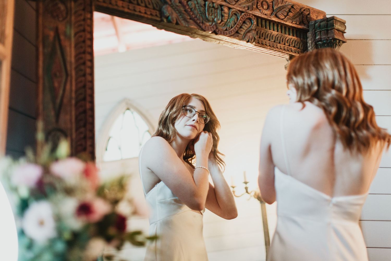 Bride in white gown adjusting earring while looking in ornate mirror, with floral bouquet in foreground.