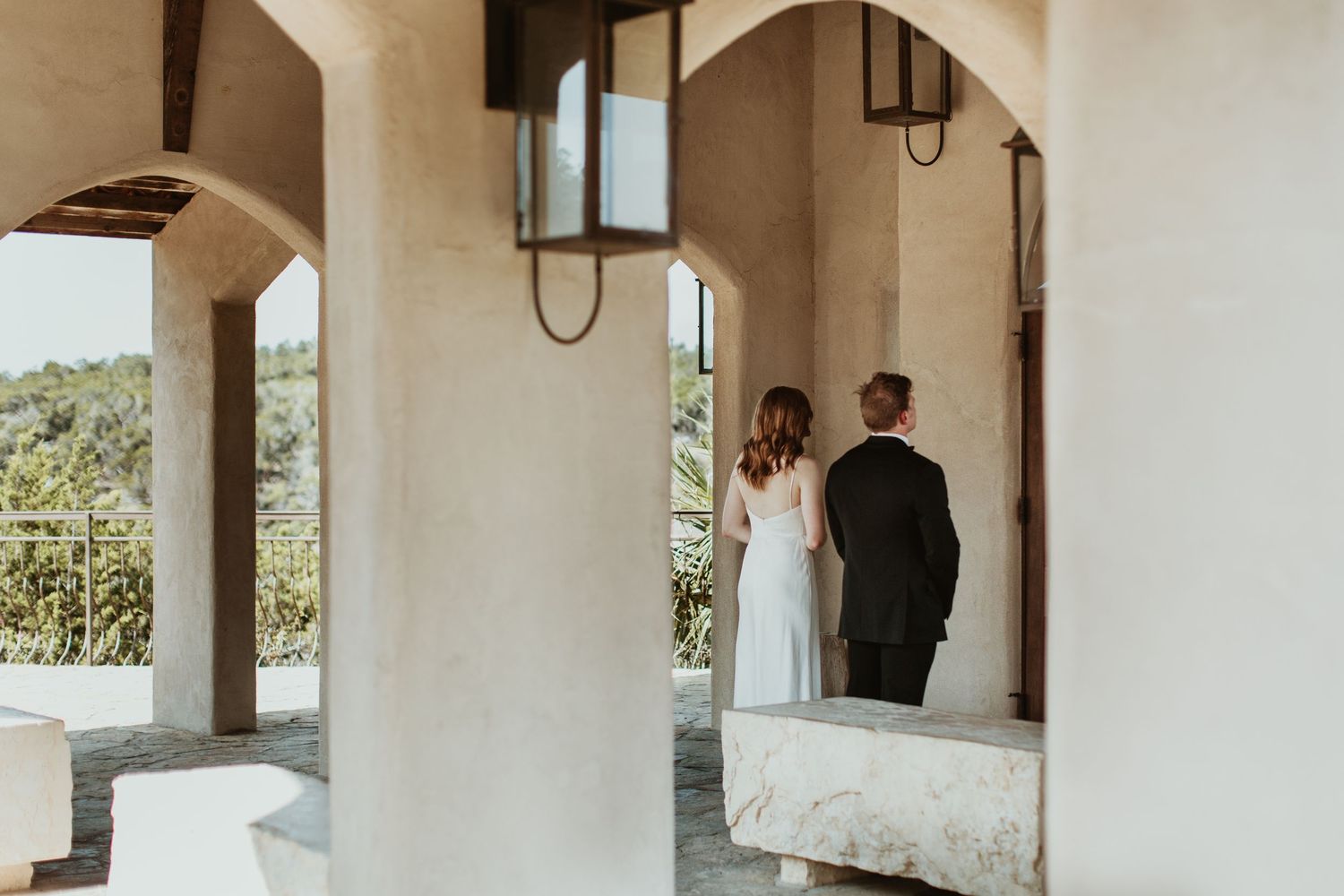 A bride and groom stand together under arched columns of an elegant venue, gazing at a scenic outdoor view.