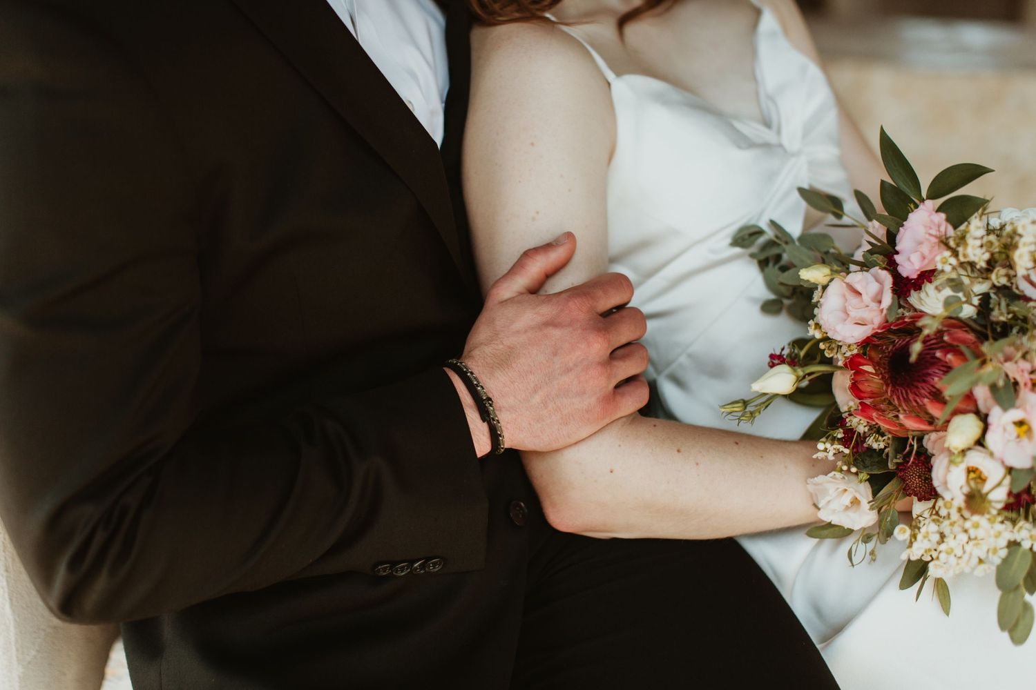 Bride and groom embracing, bride holding a lush pink and red floral bouquet, groom in dark suit.