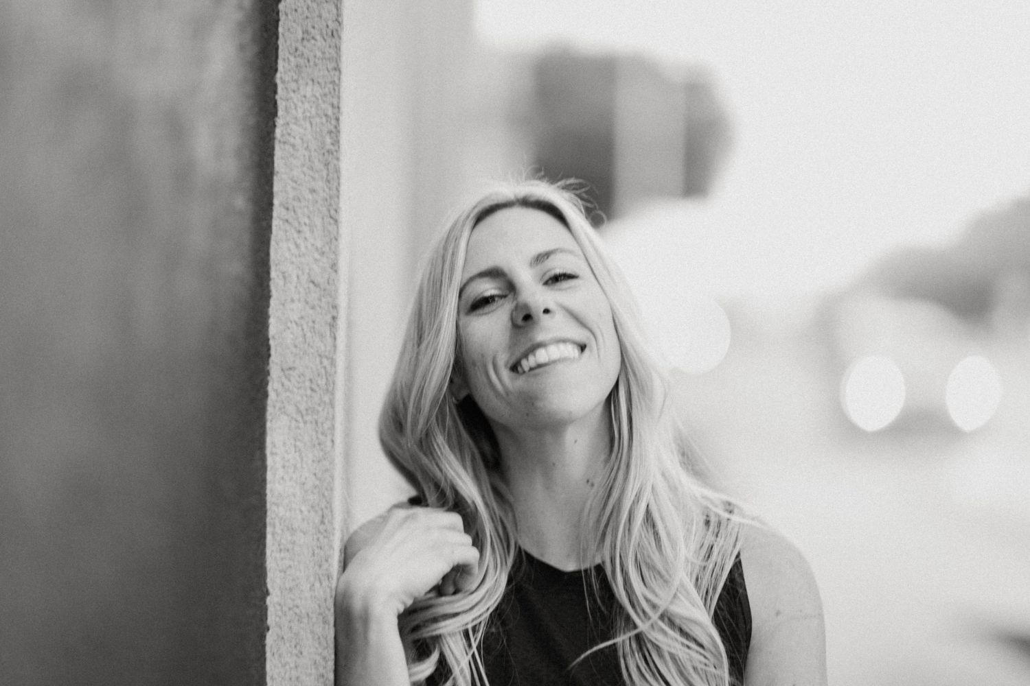 Black and white portrait of a smiling blonde woman leaning against a wall outdoors.