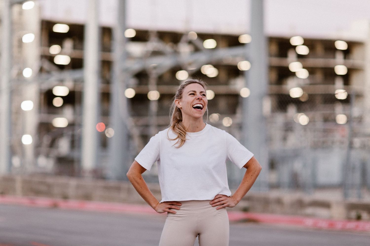 Woman in white crop top and beige leggings laughing outdoors near urban structure at dusk.