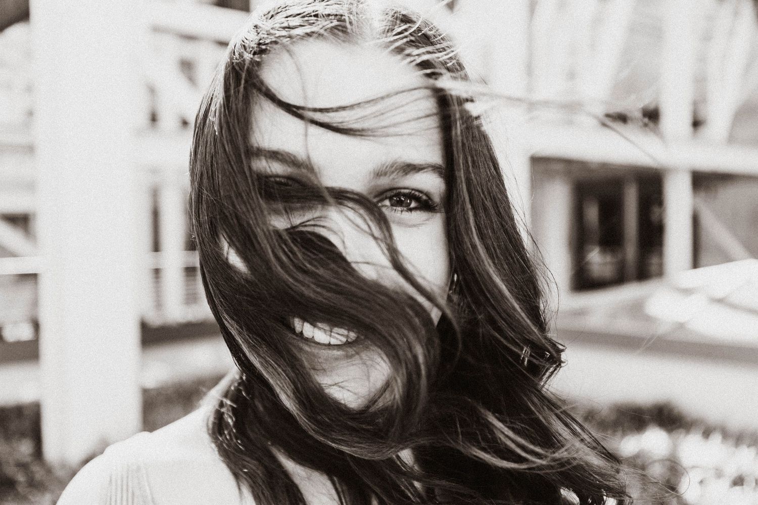 Young woman smiling with long brown hair blowing across her face in a black and white outdoor portrait.