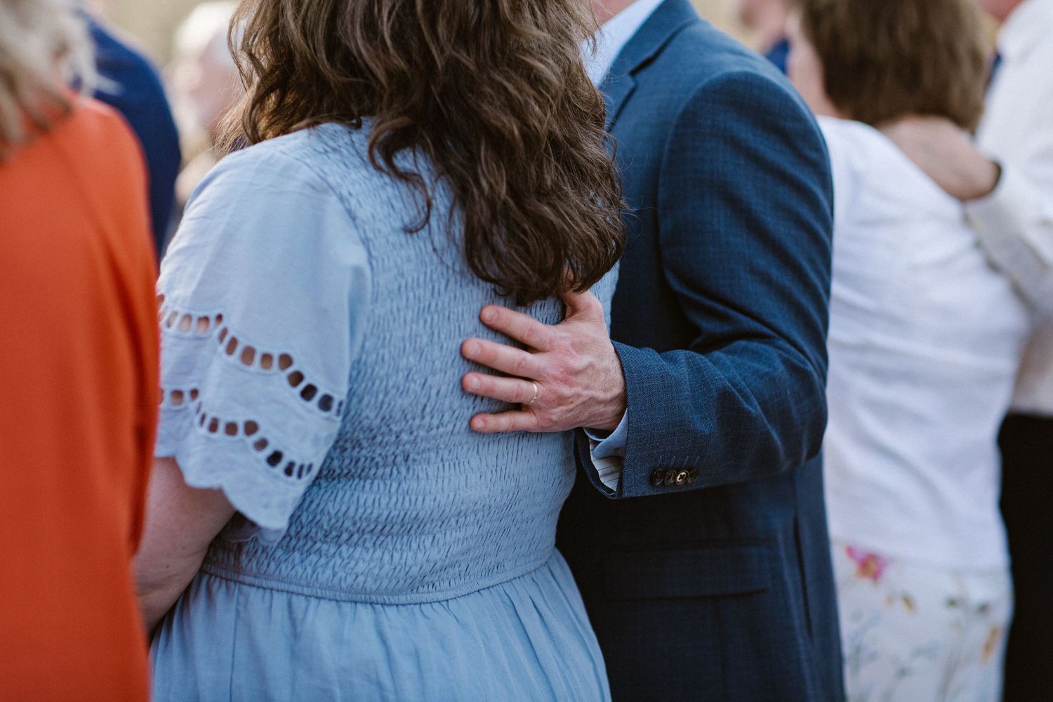 Man in navy suit places hand on womans back in light blue dress during wedding reception.