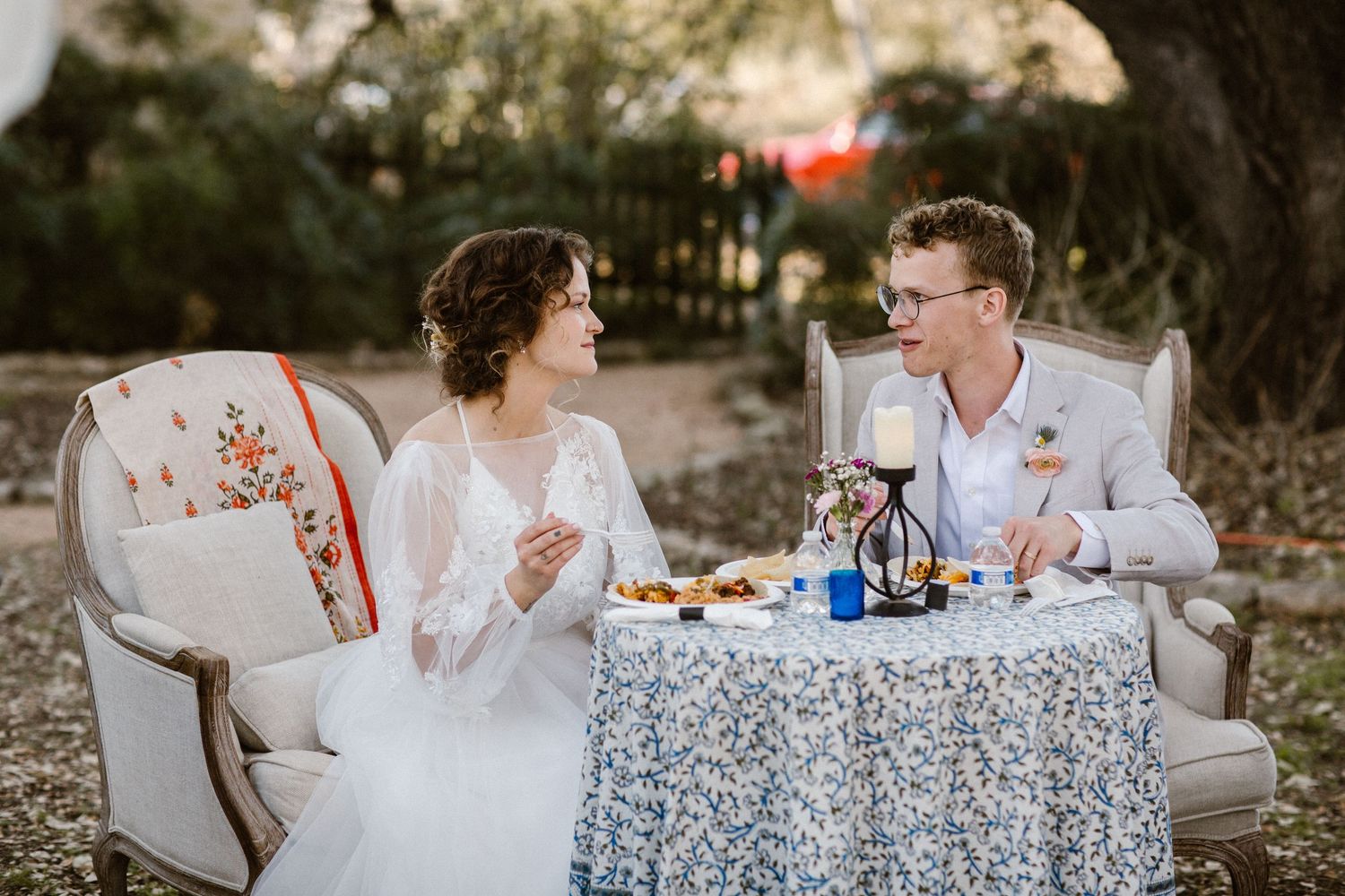 Bride and groom share a private moment seated at a small decorated table outdoors.